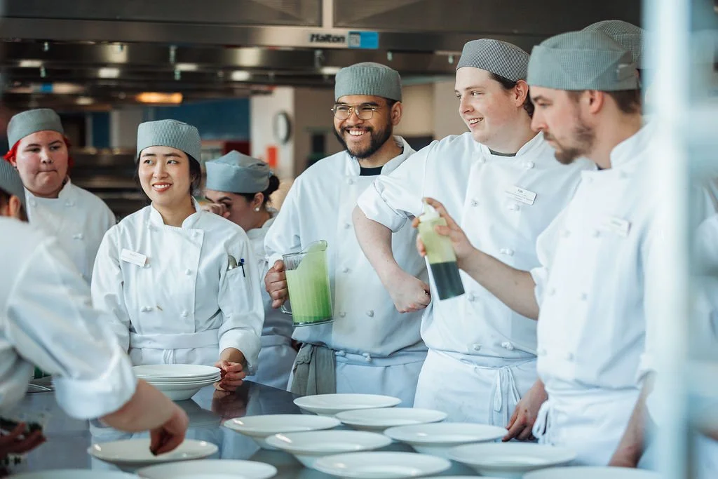 Chef instructors and students in a culinary classroom, wearing white coats and gray hats, discussing food preparation with plates and a blender of green smoothie on the counter.