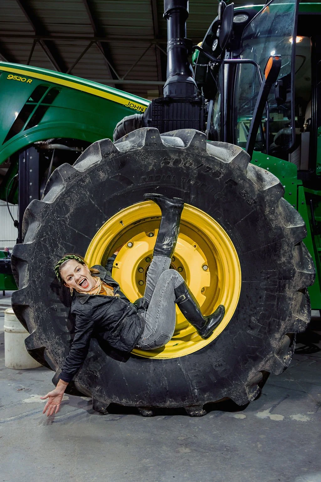 A woman is lying on a large tractor tire with a big smile, inside a warehouse or indoor farm equipment area.