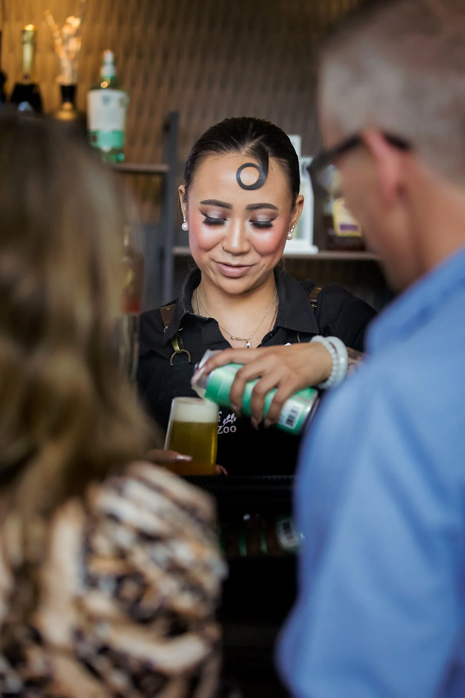 A woman with dark hair and pearl earrings serving drinks at a bar, with two blurred people in the foreground.