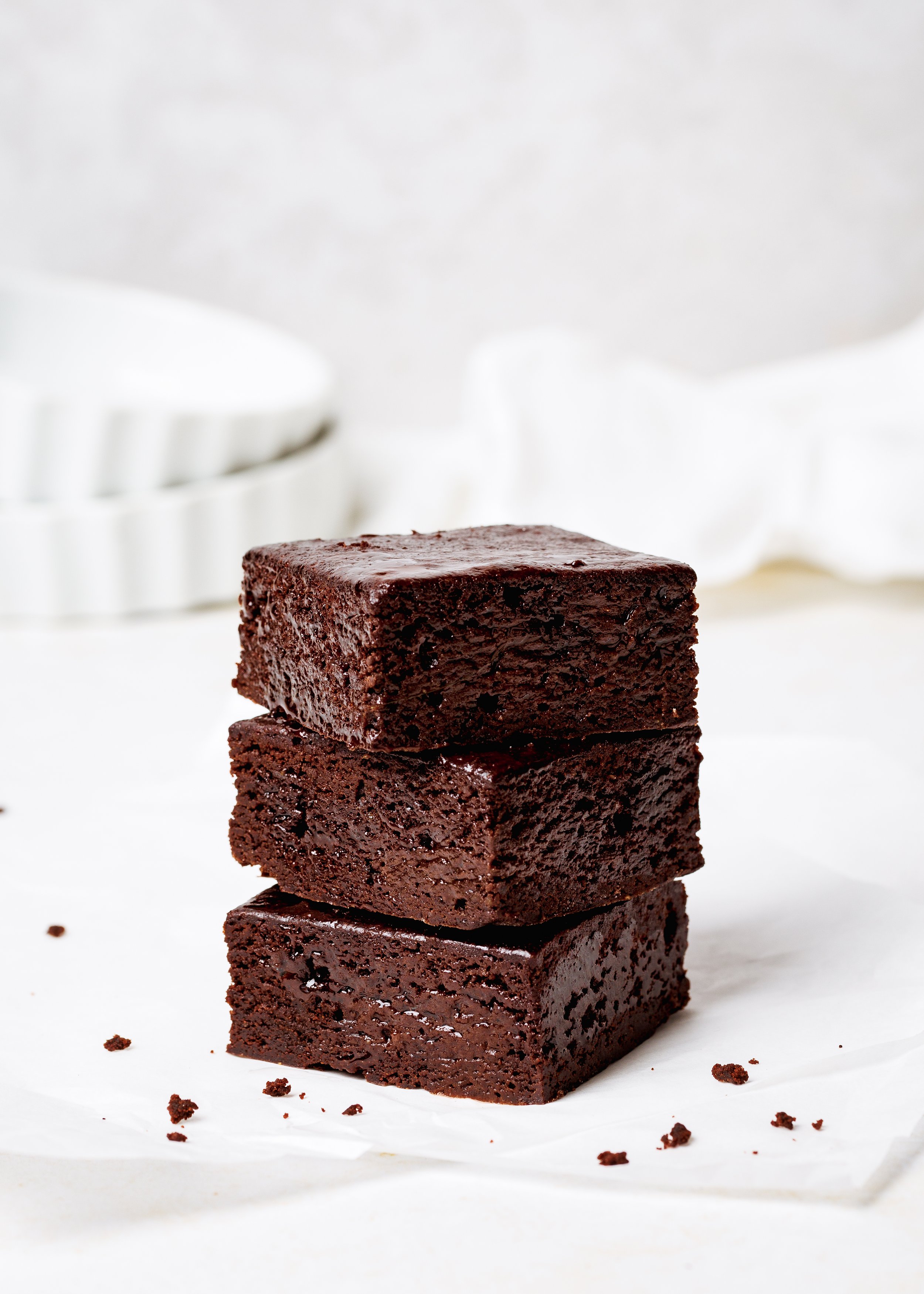 Stack of four chocolate brownies on white surface with scattered crumbs, blurred white background.