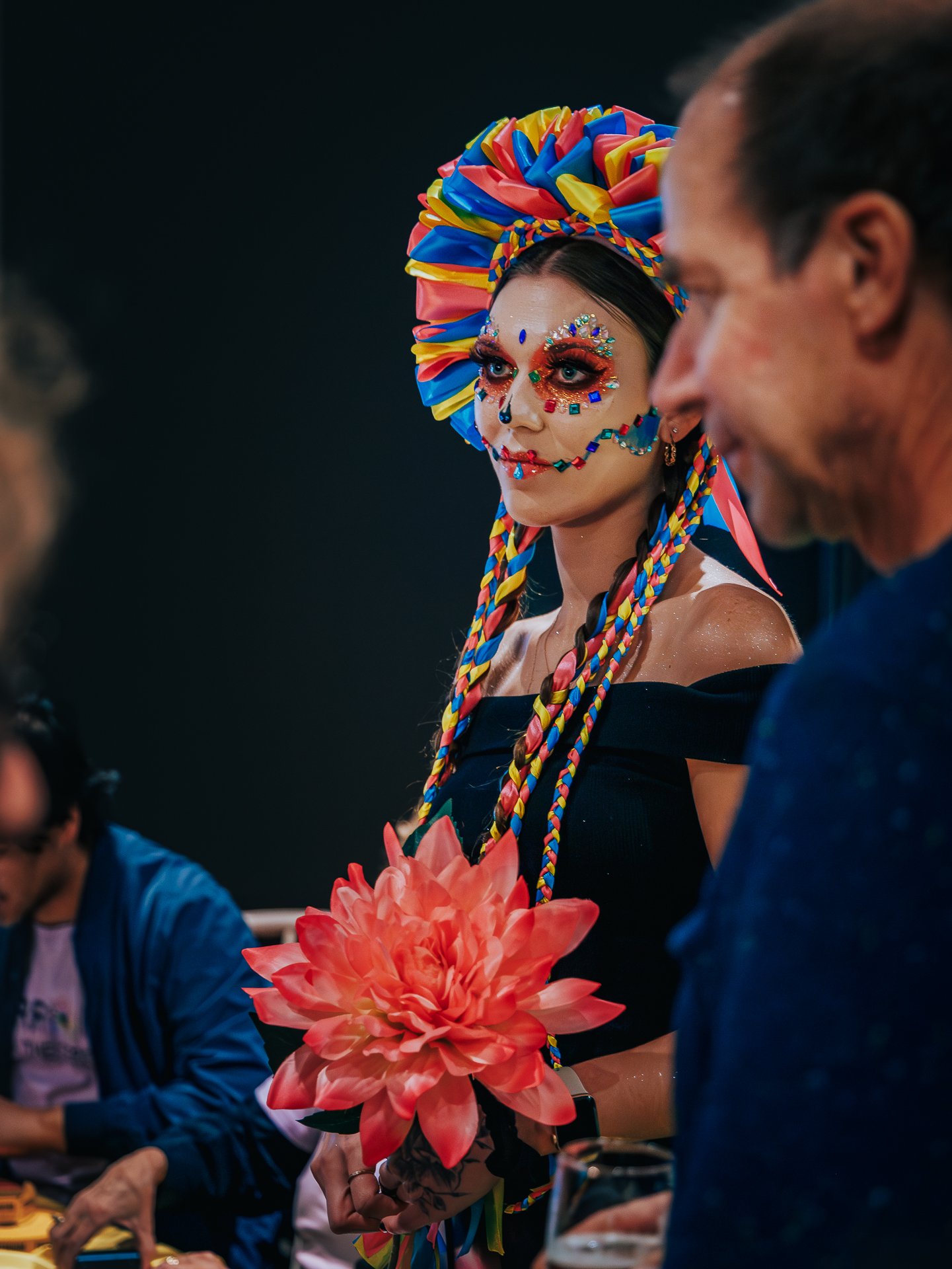 Woman dressed in colorful costume with elaborate face makeup and a large multicolored headpiece, holding a big red flower, participating in a festive event.