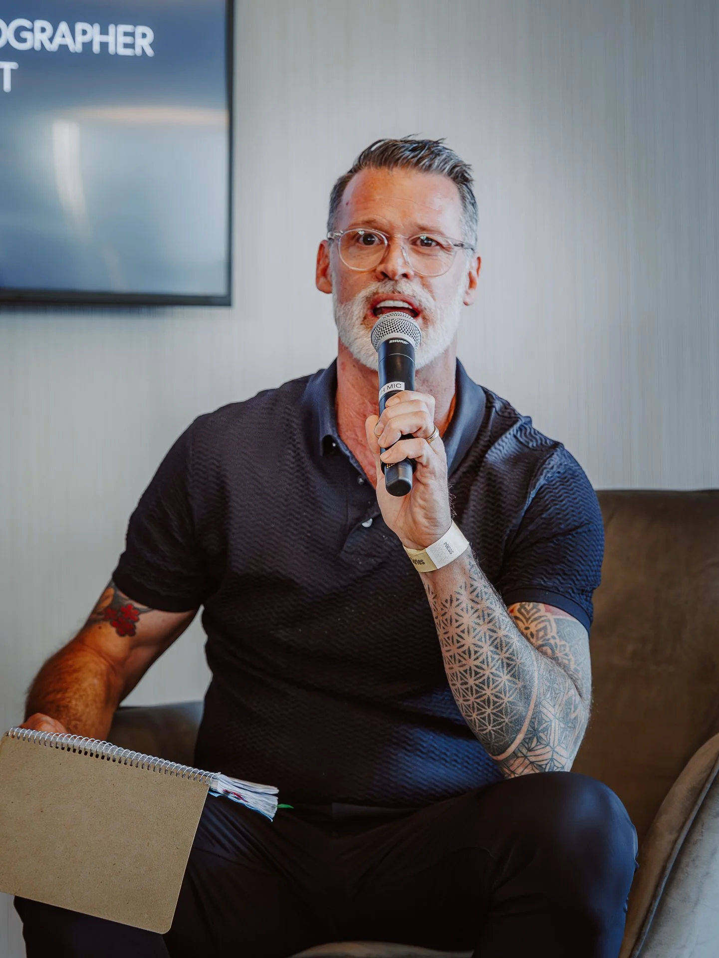 A man with glasses and tattoos on his arm speaking into a microphone during a presentation or speech, sitting at a table with a notepad and a folder.
