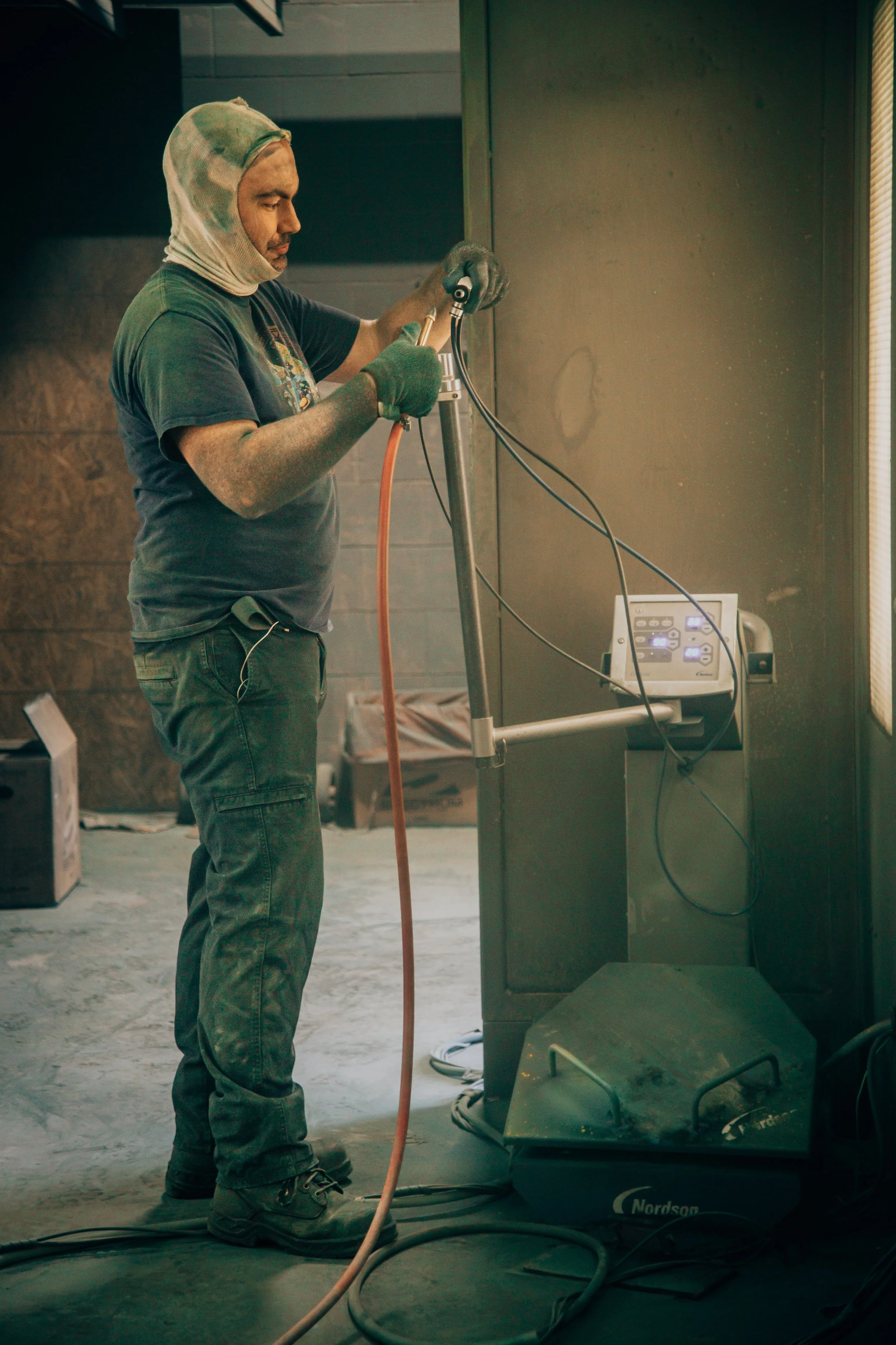 A person wearing a balaclava, gloves, and casual clothes is working with welding or cutting equipment in an industrial setting. The individual appears focused, standing beside a machine with electrical cords and a control panel, with graffiti on the 