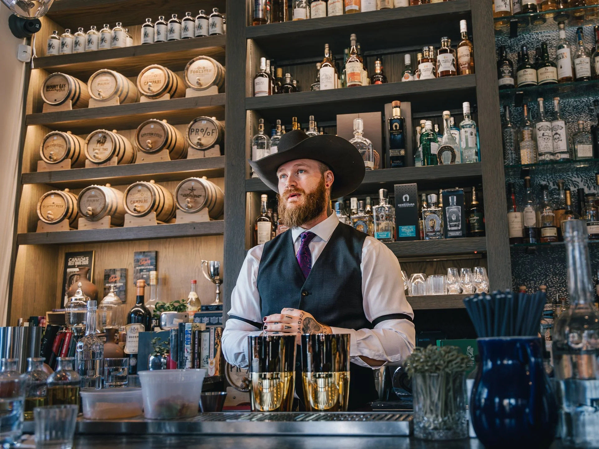 A bartender with a beard and tattoo on his hand, wearing a cowboy hat and dressed in a vest and white shirt, standing behind a bar with bottles of liquor, glasses, and bar tools on display in a bar or lounge setting.