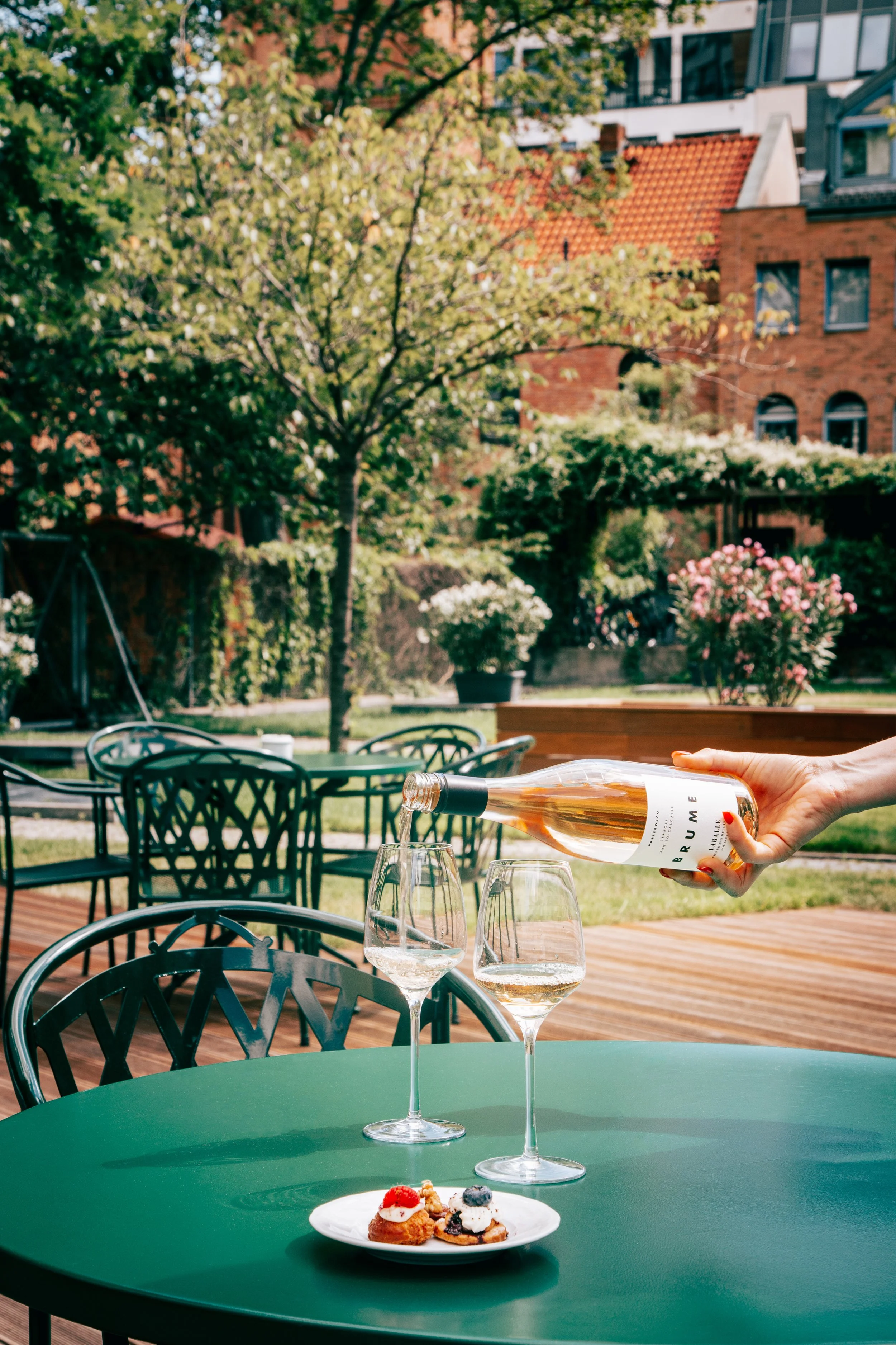 A person is pouring wine into a glass on a green outdoor table. There are two glasses of wine, a dessert plate with pastries topped with berries, and a green table with black chairs in the background. The setting is at a patio or garden area with tre