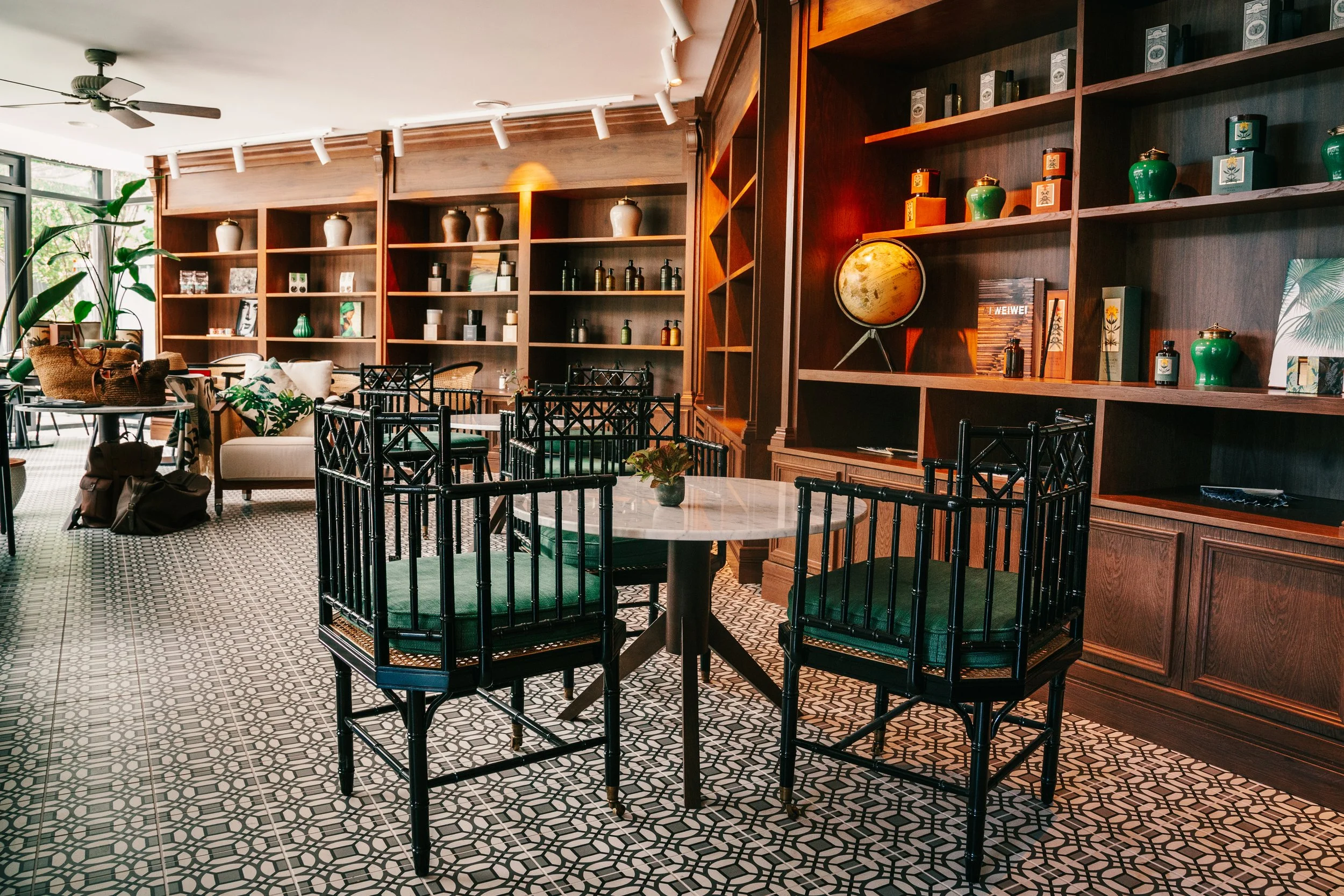 Interior of a cozy cafe or restaurant with wooden bookshelves filled with pottery and decorative items, black chairs around a marble table, patterned tile flooring, and large windows with plants.