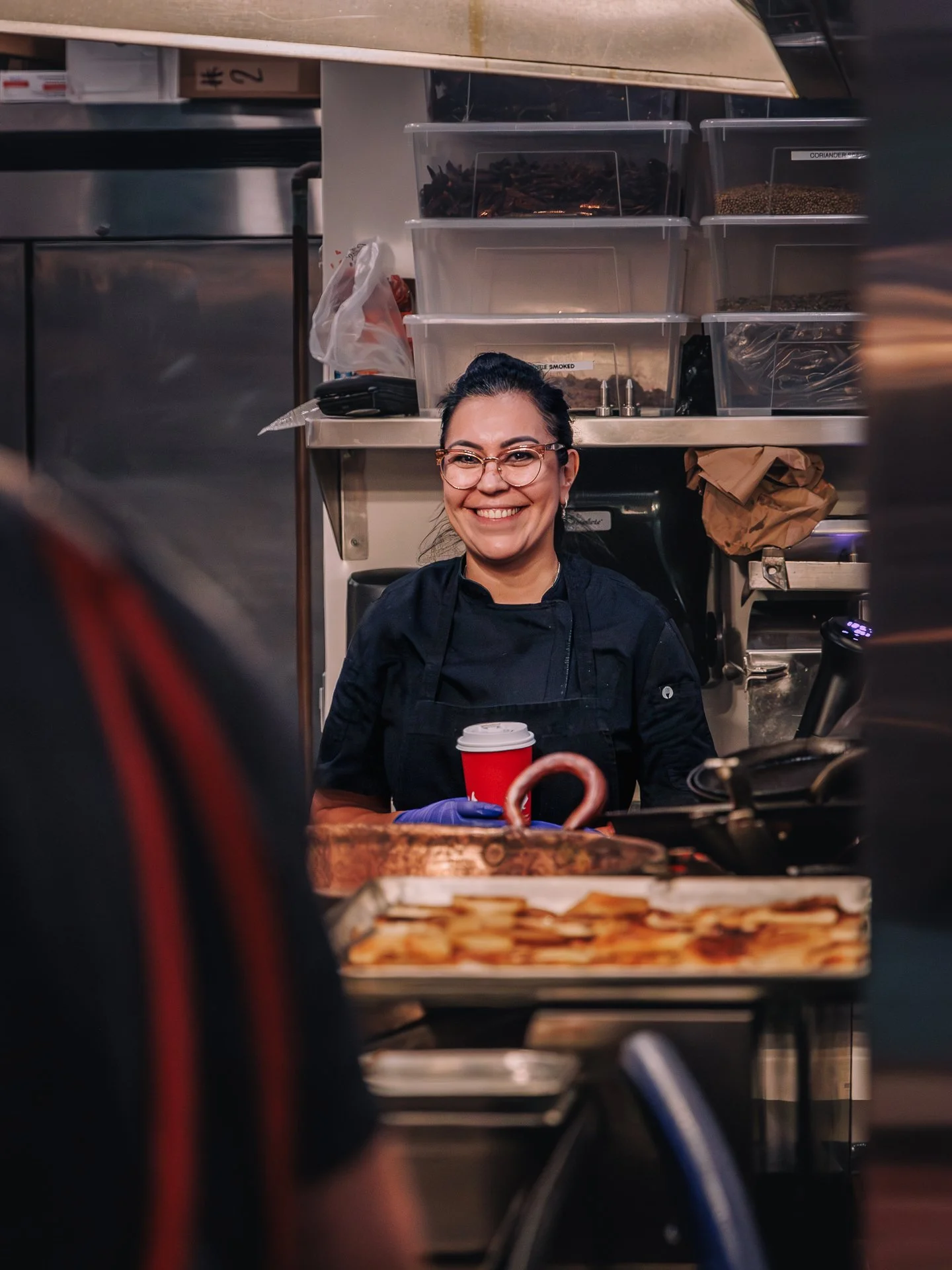A smiling woman with glasses and dark hair tied back, wearing a black uniform, holding a red disposable coffee cup in a kitchen or food service area.