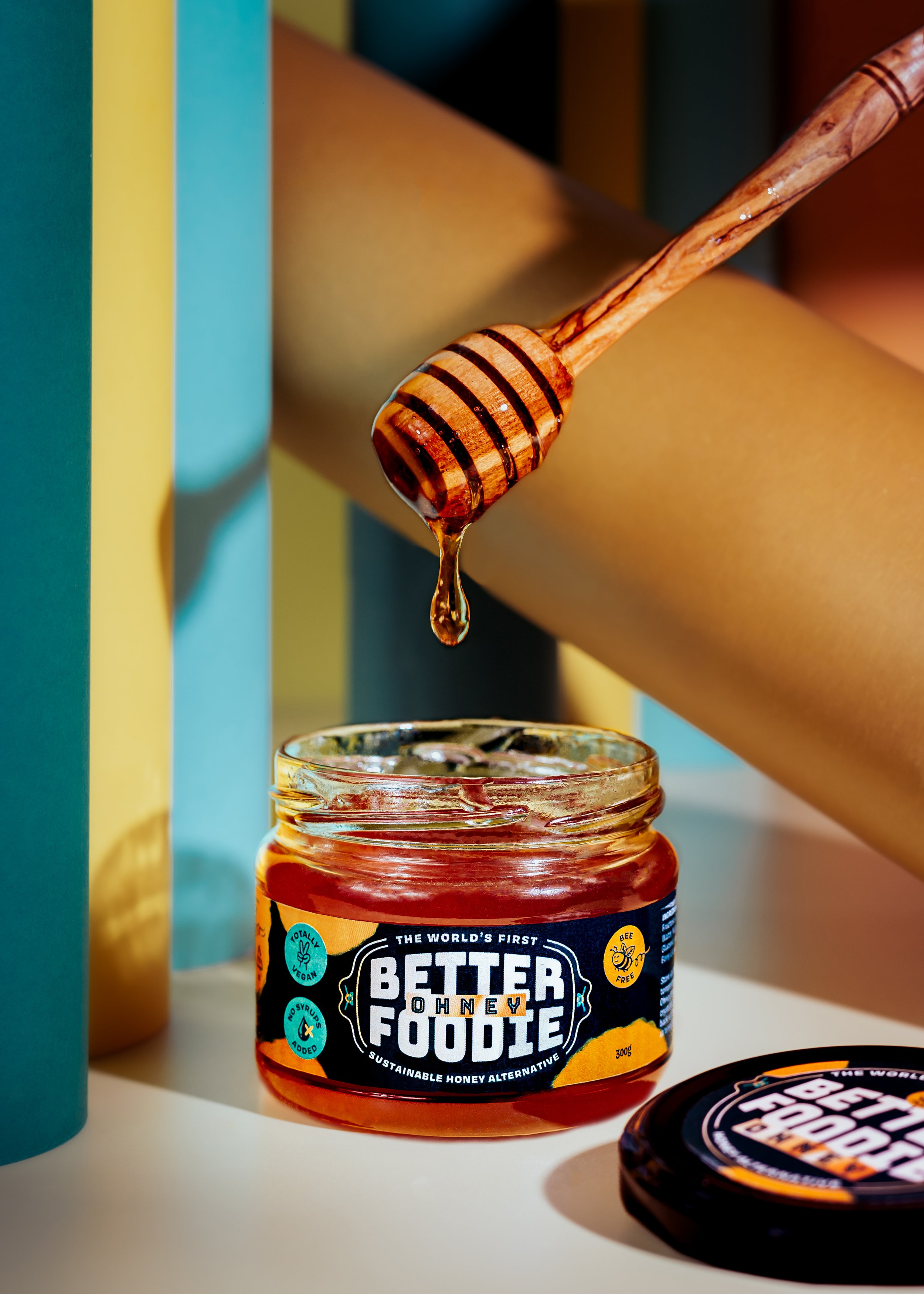 A jar of honey labeled 'Better for You Honey', with a honey dipper lifting honey from the jar. The label indicates it is a sustainable, vegan, and no-sugar-added alternative. In the background, part of a blue container and a yellow object, possibly a