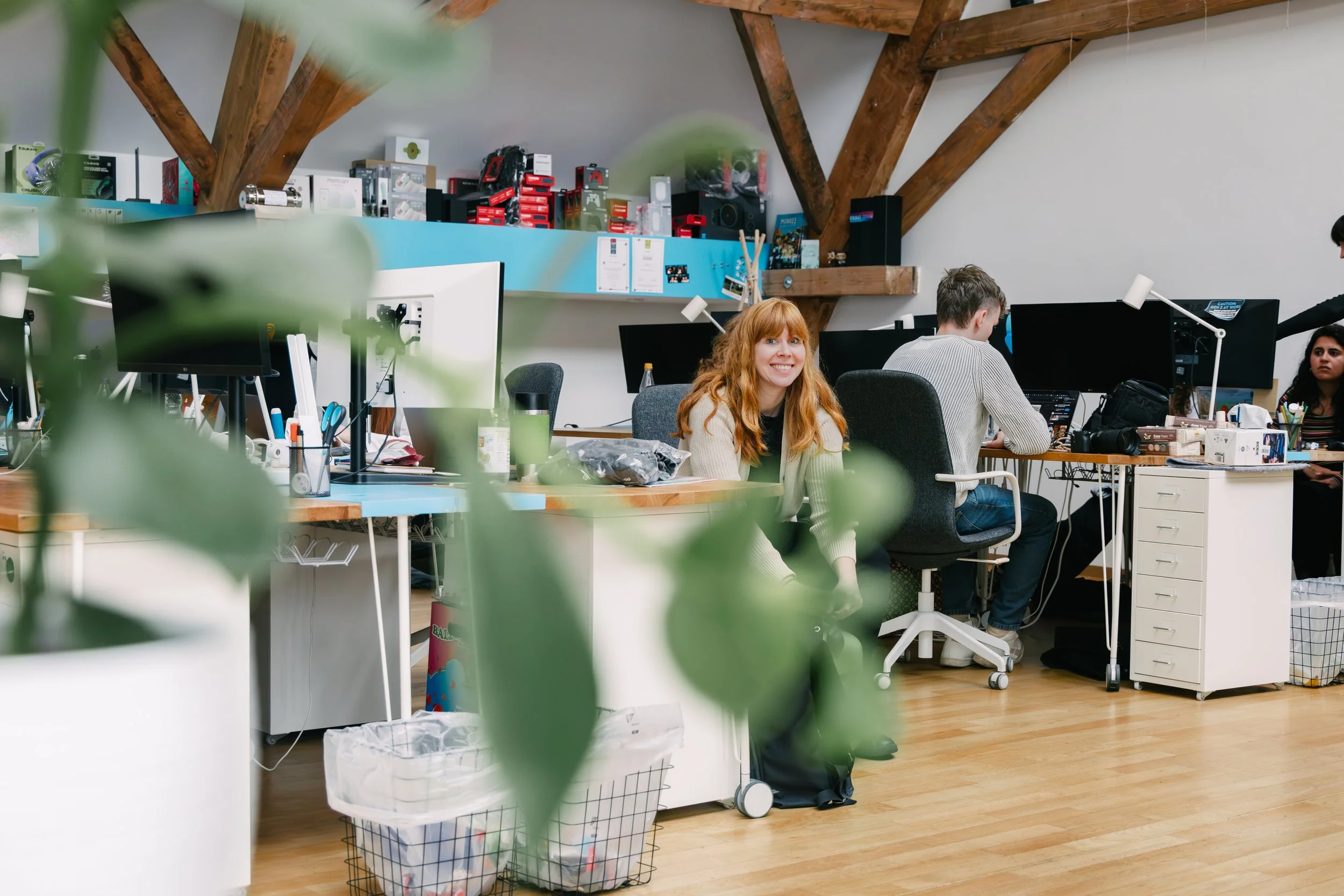 Office workspace with people working at desks, computer monitors, shelves with boxes, and a person smiling at the camera.