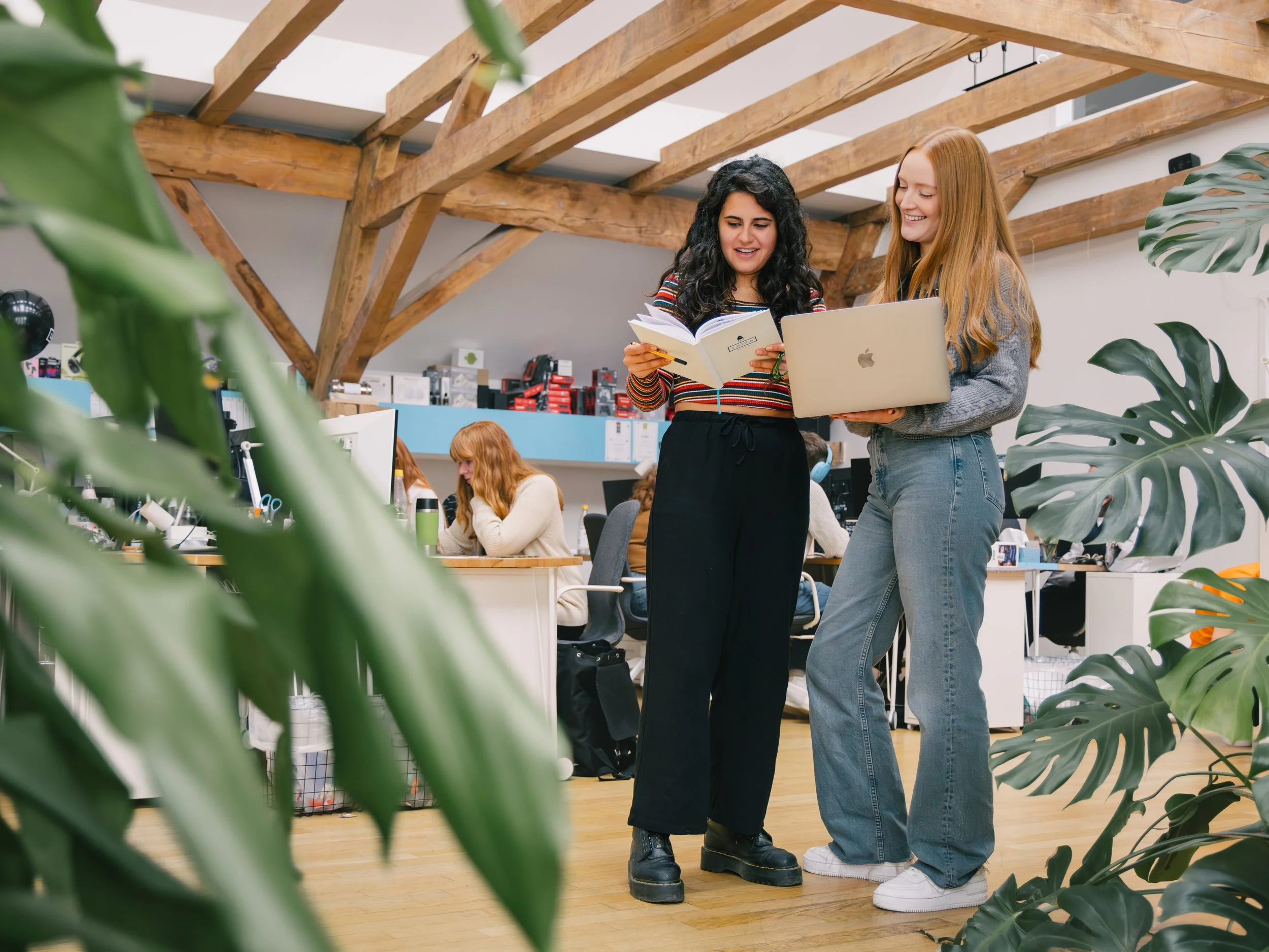 Two women are standing in an office, looking at a notebook and a laptop while smiling. The office has a wooden beam ceiling and plants in the foreground, with other people working at desks in the background.