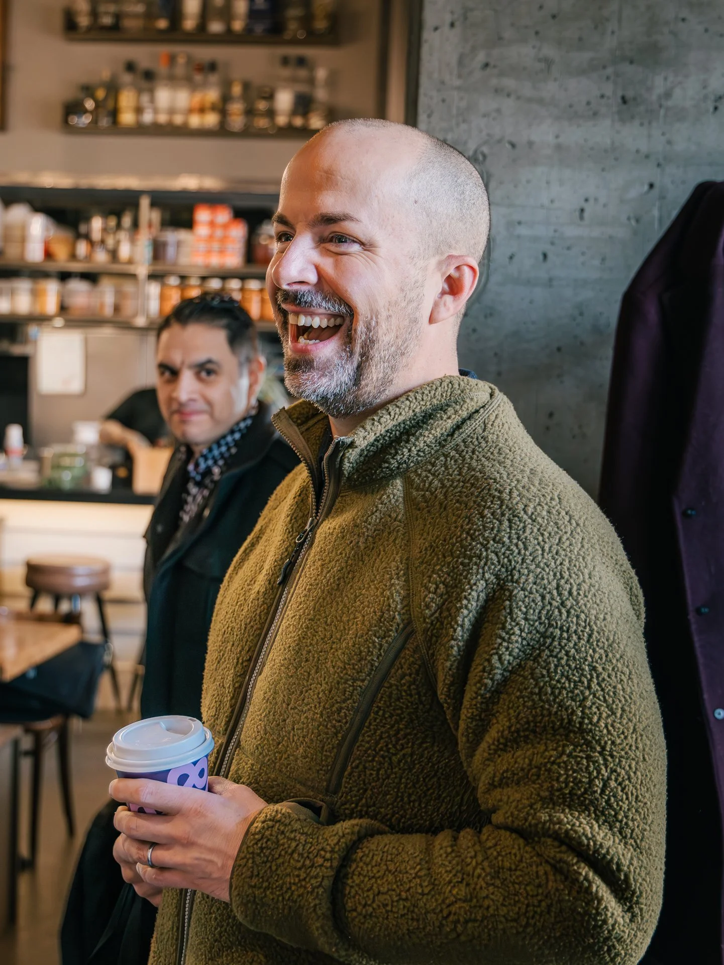 Man with a beard smiling while holding a coffee cup in a casual indoor setting, with another man blurred in the background, shelves with jars and condiments, and a grey concrete wall.
