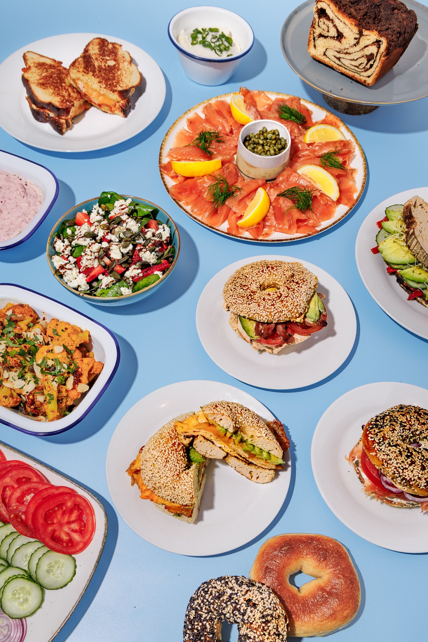 A variety of foods on a blue table, including bagels, sandwiches, a smoked salmon platter with lemon wedges and capers, a bowl of salad with vegetables and crumbled cheese, a bowl of cream with green onions, a slice of marble cake, a fruit salad with
