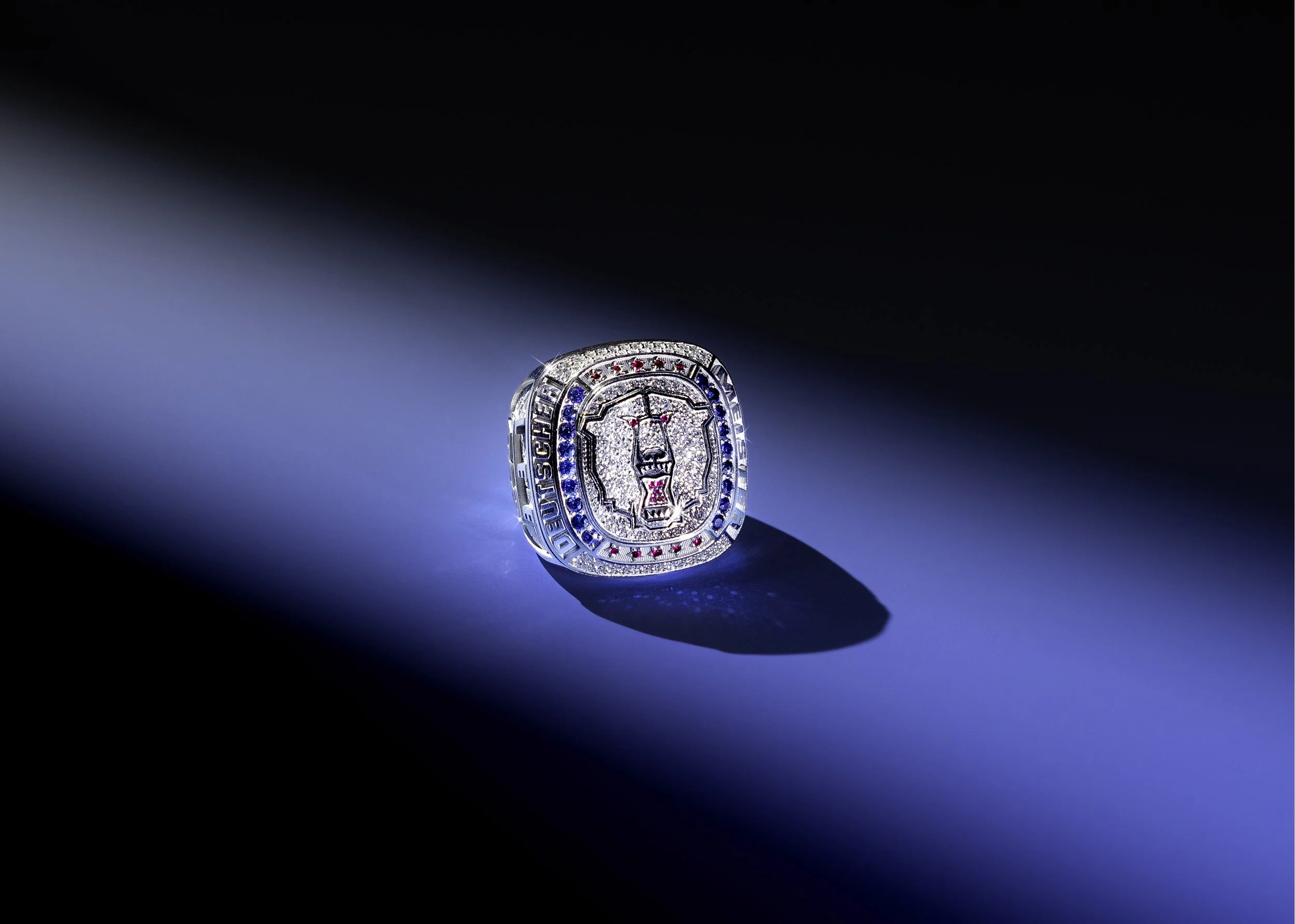 A close-up of a luxury jewelry ring with a lion's face design, encrusted with diamonds, blue and red gemstones, illuminated against a dark background.