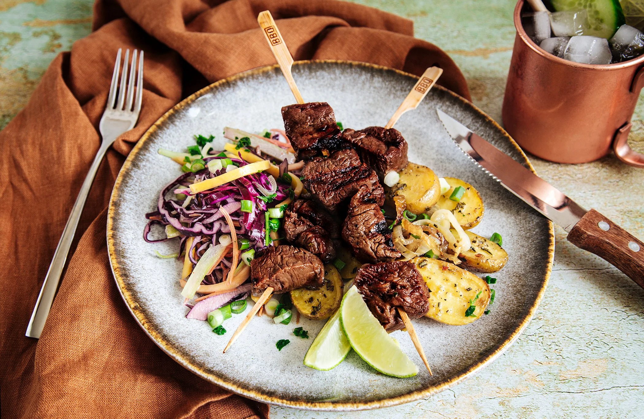 Close-up of a plate with grilled steak skewers, roasted potatoes, and colorful salad with cabbage, sliced vegetables, and chopped green onions, garnished with lime wedges.