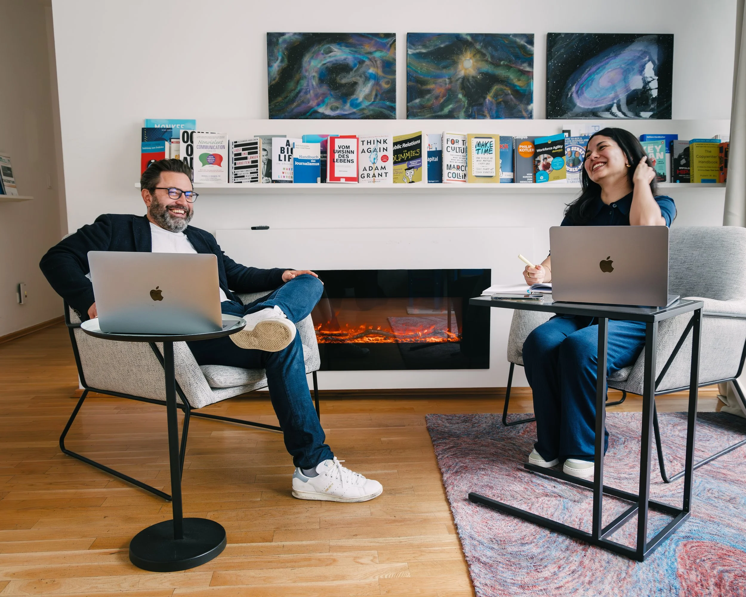 Two people sitting in a room with a fireplace and a bookshelf, laughing and smiling at each other, with paintings of the galaxy above the bookshelf.