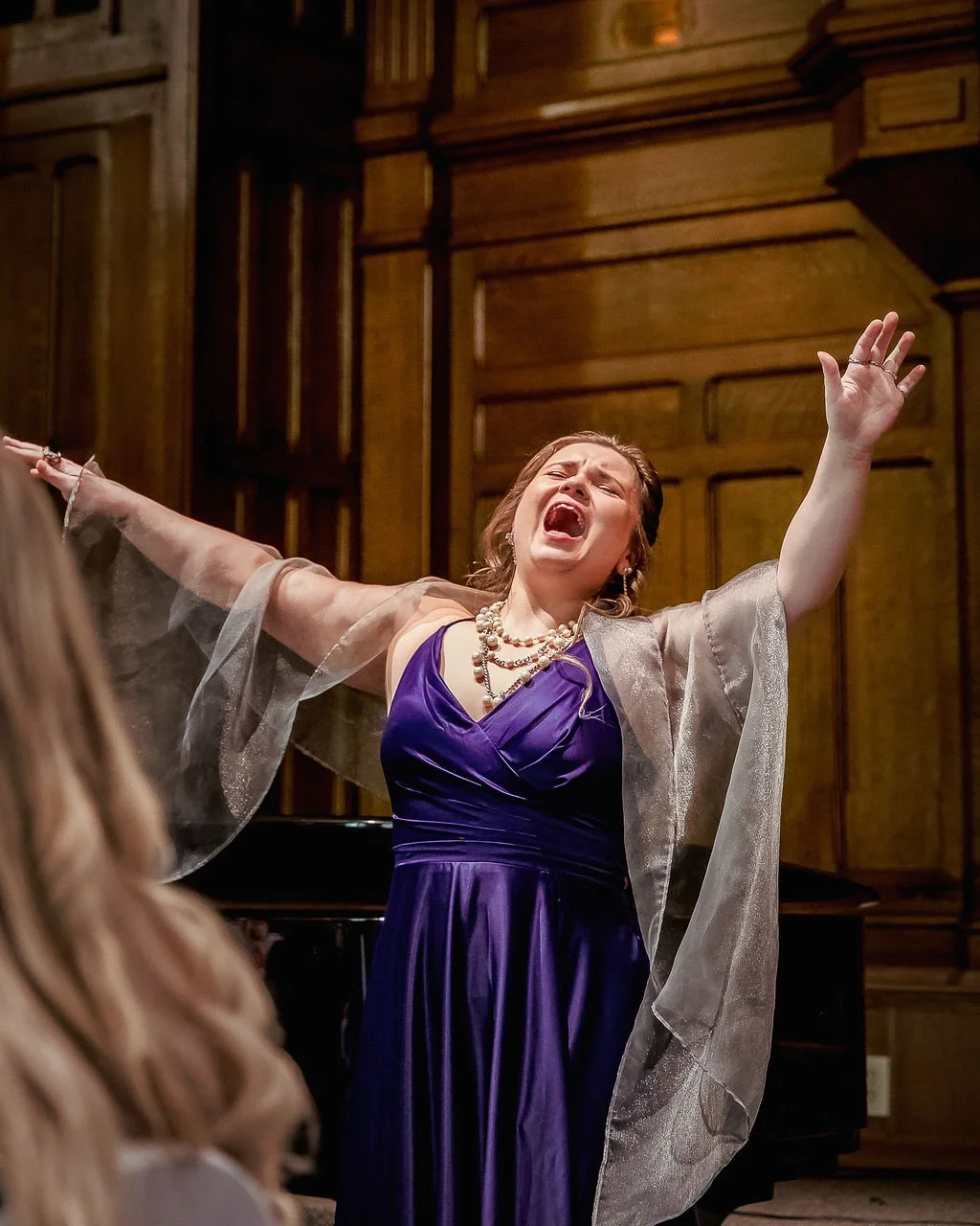 A woman in a purple dress and pearl necklace passionately singing with arms outstretched in a wood-paneled room.