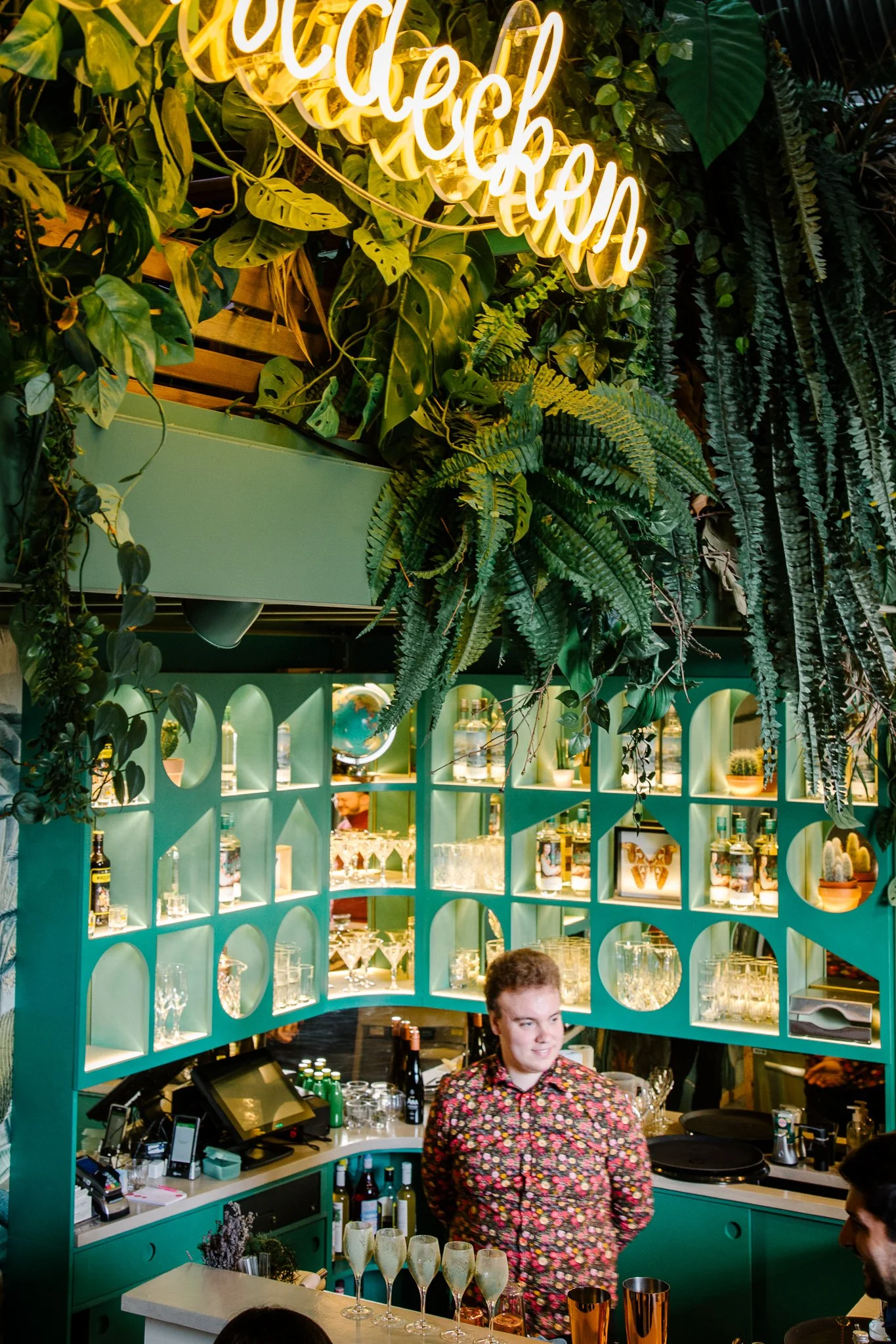 A bartender stands behind a bar counter decorated with green plants and a teal-colored partition with oval cutouts. The bartender is smiling and wearing a floral shirt. On the bar are champagne glasses, bottles, and bar tools. A neon sign above reads