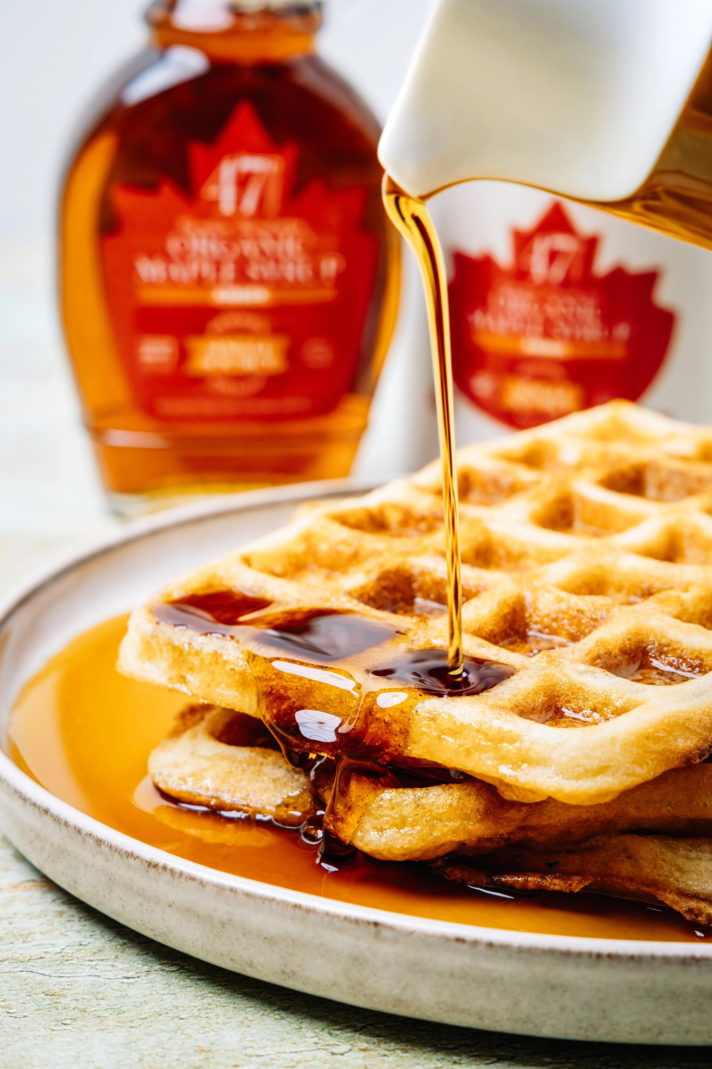 Close-up of a waffle with syrup being poured onto it from a bottle, with a jar of syrup in the background.
