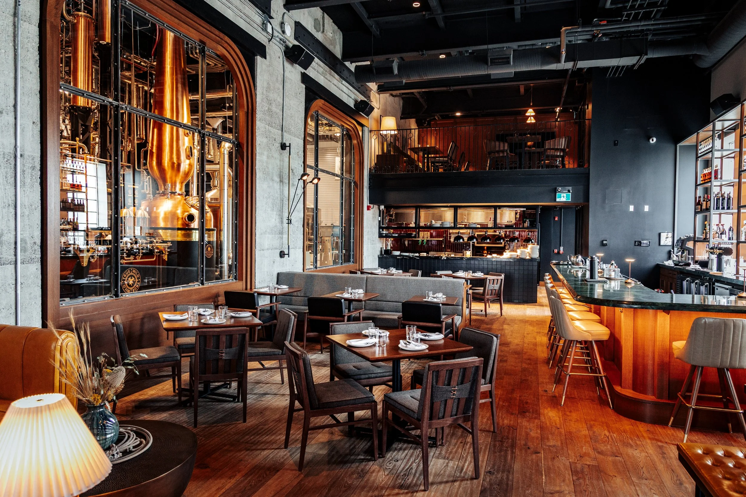 Interior of a modern restaurant with wooden floors, black walls, and contemporary furniture. Large copper distillation apparatus is visible through glass windows, indicating a distillery setting.