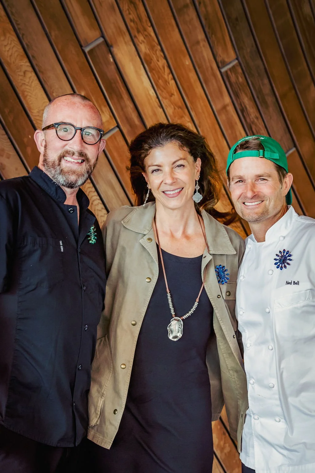 Three people smiling, standing close together against a wooden wall. From left to right: a man with glasses and a beard wearing a black button-up shirt, a woman with wavy dark hair wearing earrings, a necklace, and a beige jacket, and a man with shor