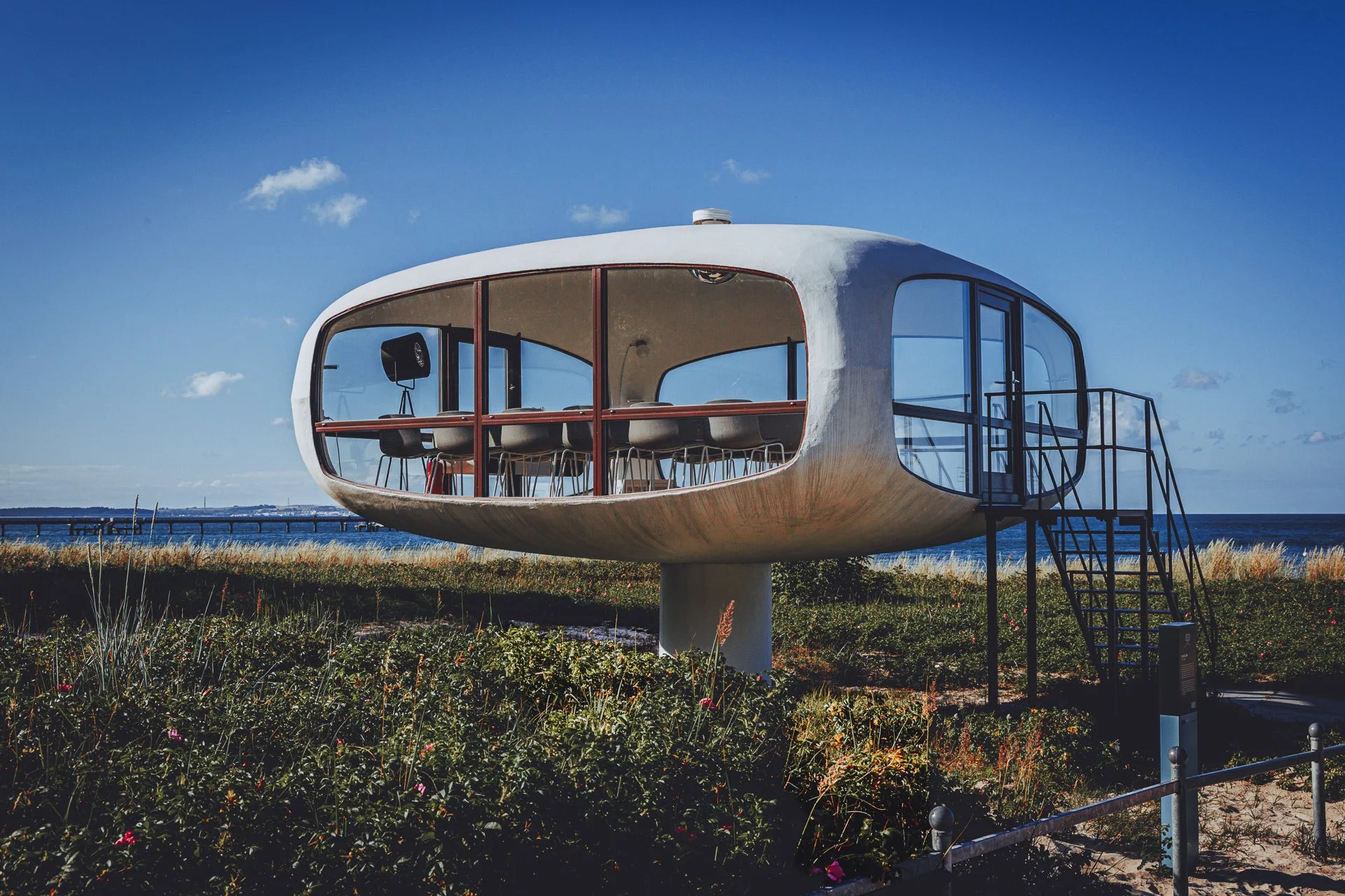 A futuristic round building with large glass windows and an external staircase, situated in a grassy area near the water under a blue sky with sparse clouds.