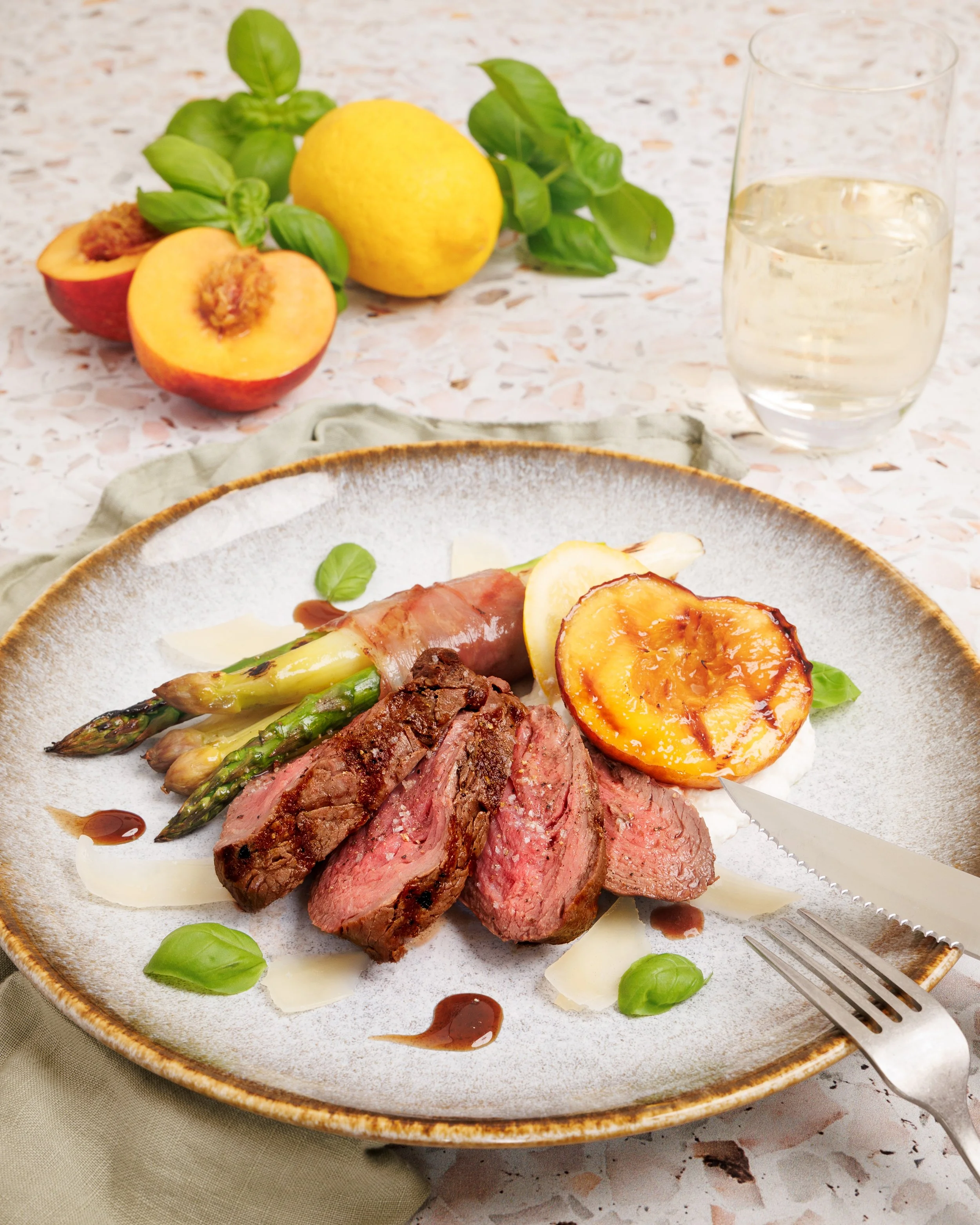 A plate of medium-rare steak with grilled asparagus, sliced roasted potato, and fresh basil leaves on a speckled ceramic plate, with a fork on the side. In the background, there are two glasses of white wine, a lemon, peaches, and basil leaves on a m