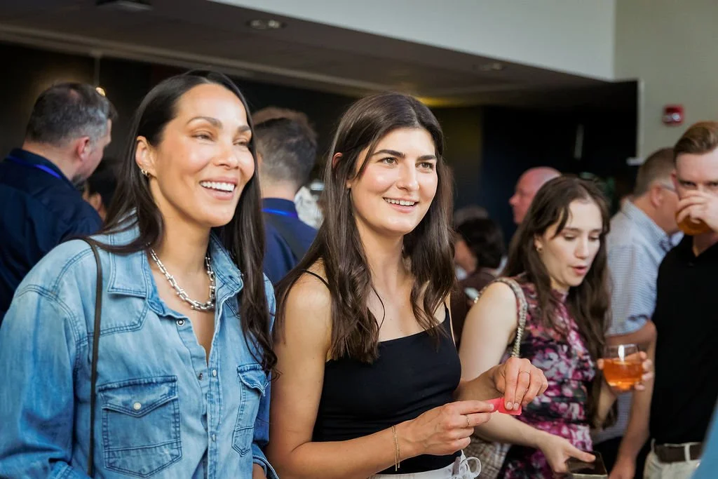Two women smiling at a social event, one in a denim jacket and the other in a black sleeveless top, among a crowd of people