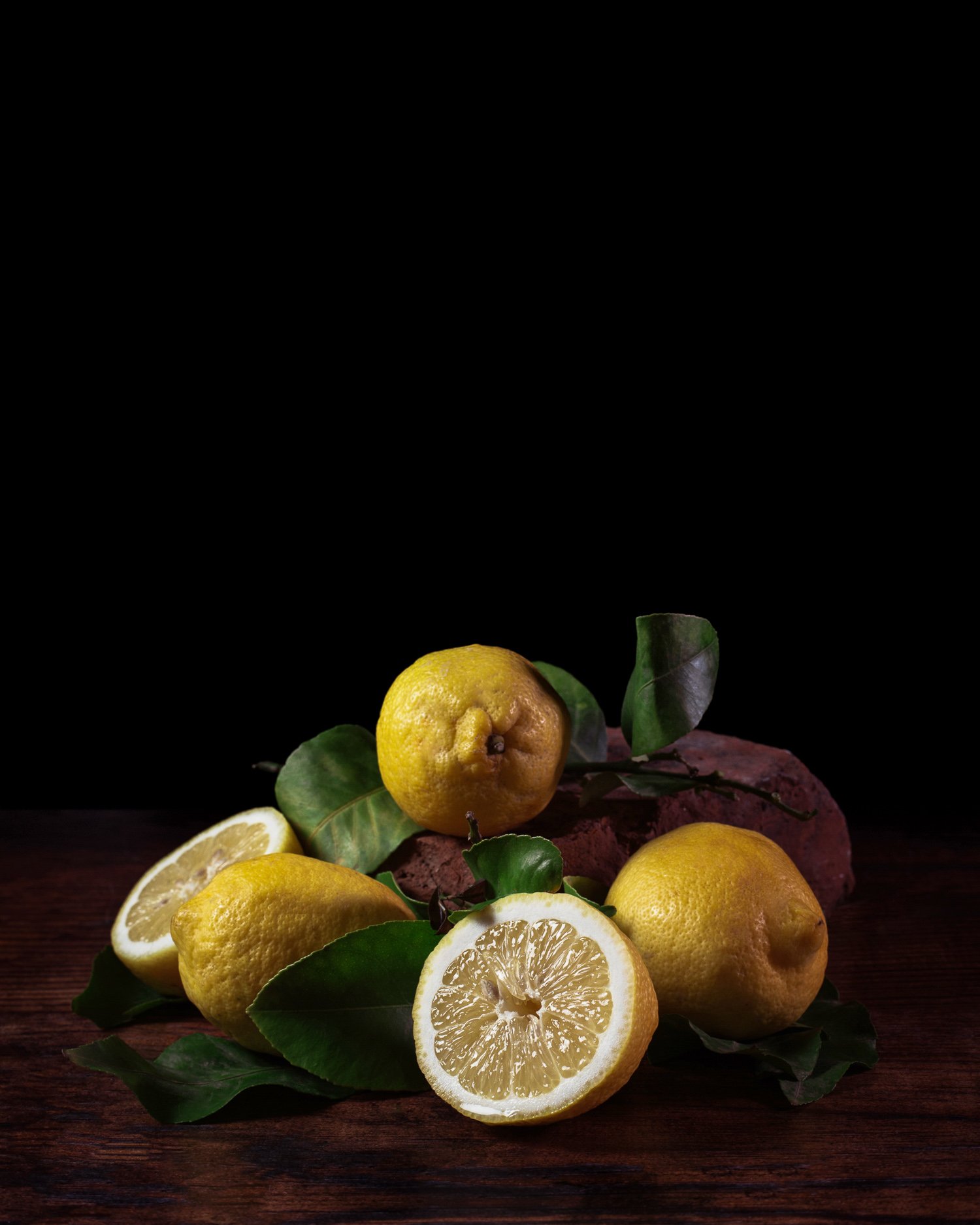 A still life composition of lemons, lemon slices, green leaves, and a wooden surface against a black background.
