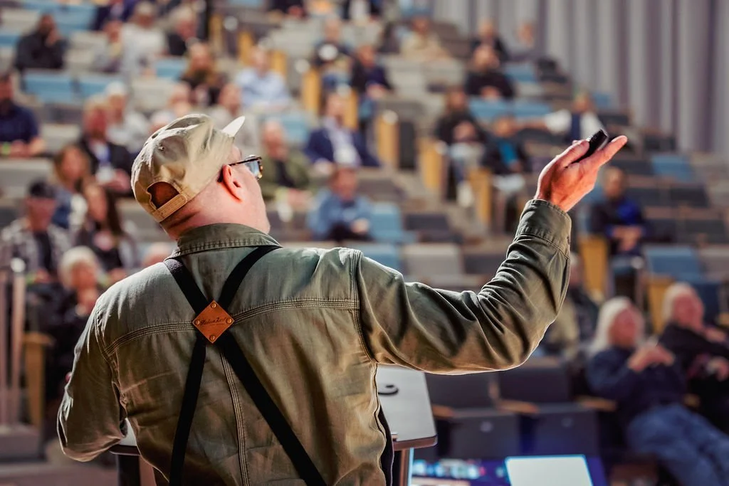 A man in a beige cap and jacket with suspenders is giving a presentation to an audience in an auditorium.