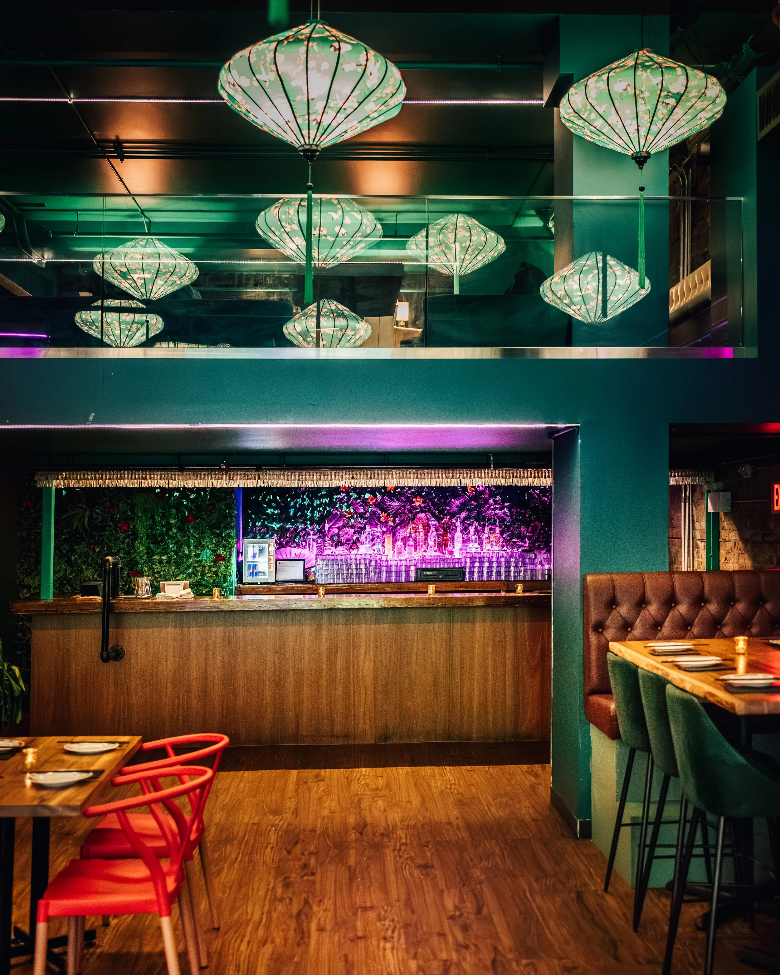 Interior of a restaurant with hanging ceiling lights, tables with chairs, and a bar area with purple lighting and a floral wall in the background.