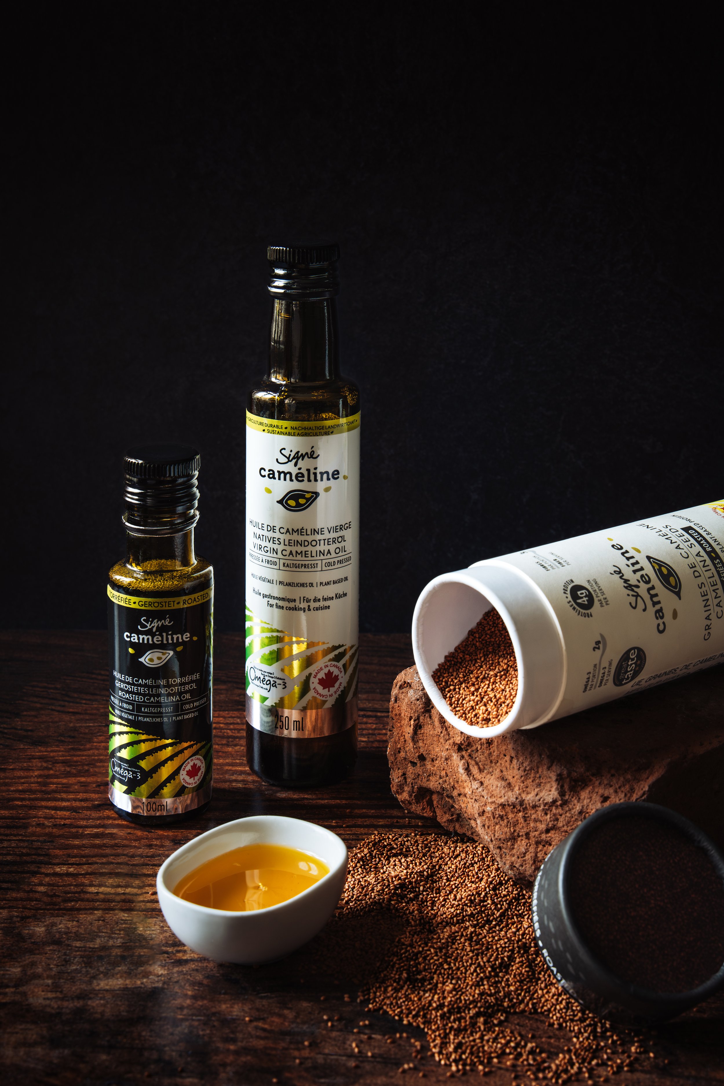 Image of bottles of organic camelina oil, a small white bowl of golden oil, a rust-colored rock, and scattered black and brown seeds on a wooden surface.