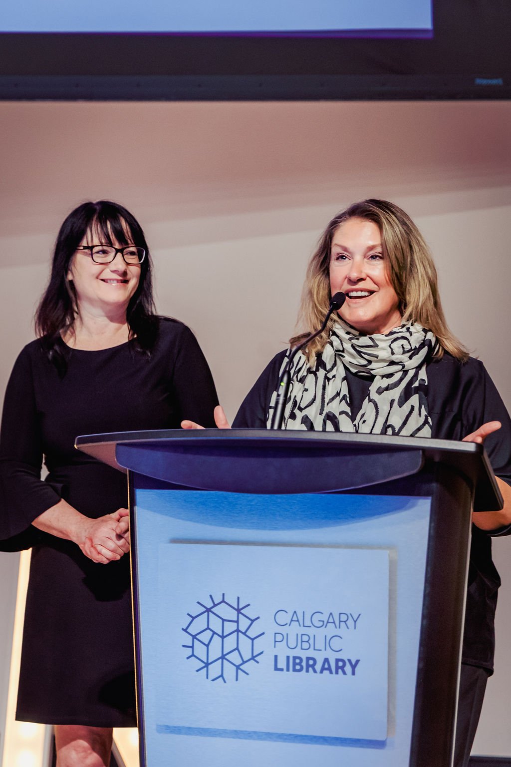 Two women standing behind a podium with a Calgary Public Library logo. One woman is speaking at a microphone, and the other is smiling.