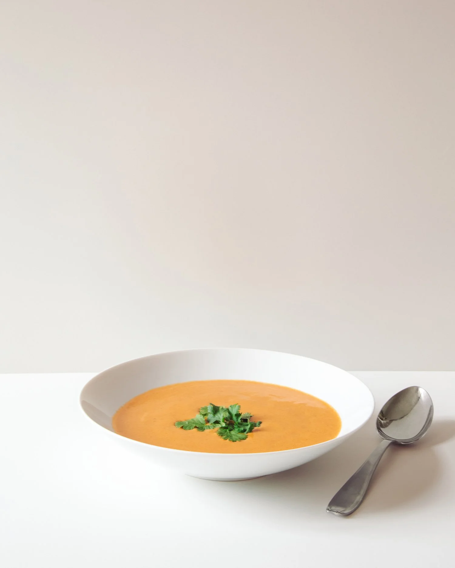A bowl of orange-colored soup garnished with cilantro leaves, with a spoon resting beside it on a white surface.