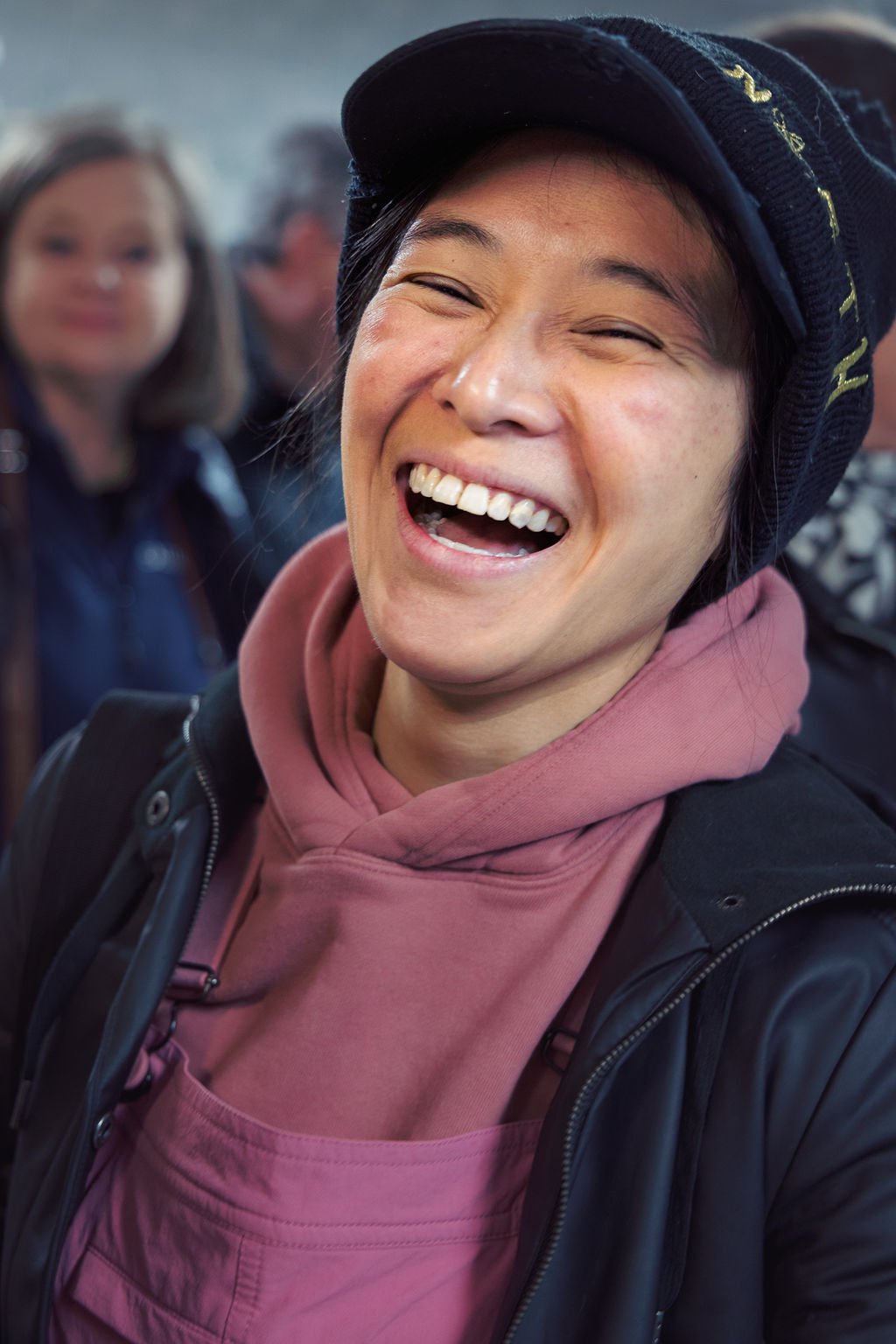 A woman laughing with eyes closed, wearing a pink hoodie, black jacket, and a dark cap, with blurred people in the background.