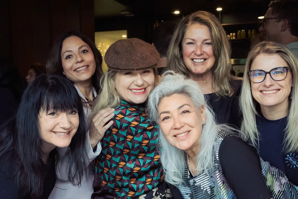 Group of six women smiling and posing for a photo at a social gathering in a coffee shop or restaurant.