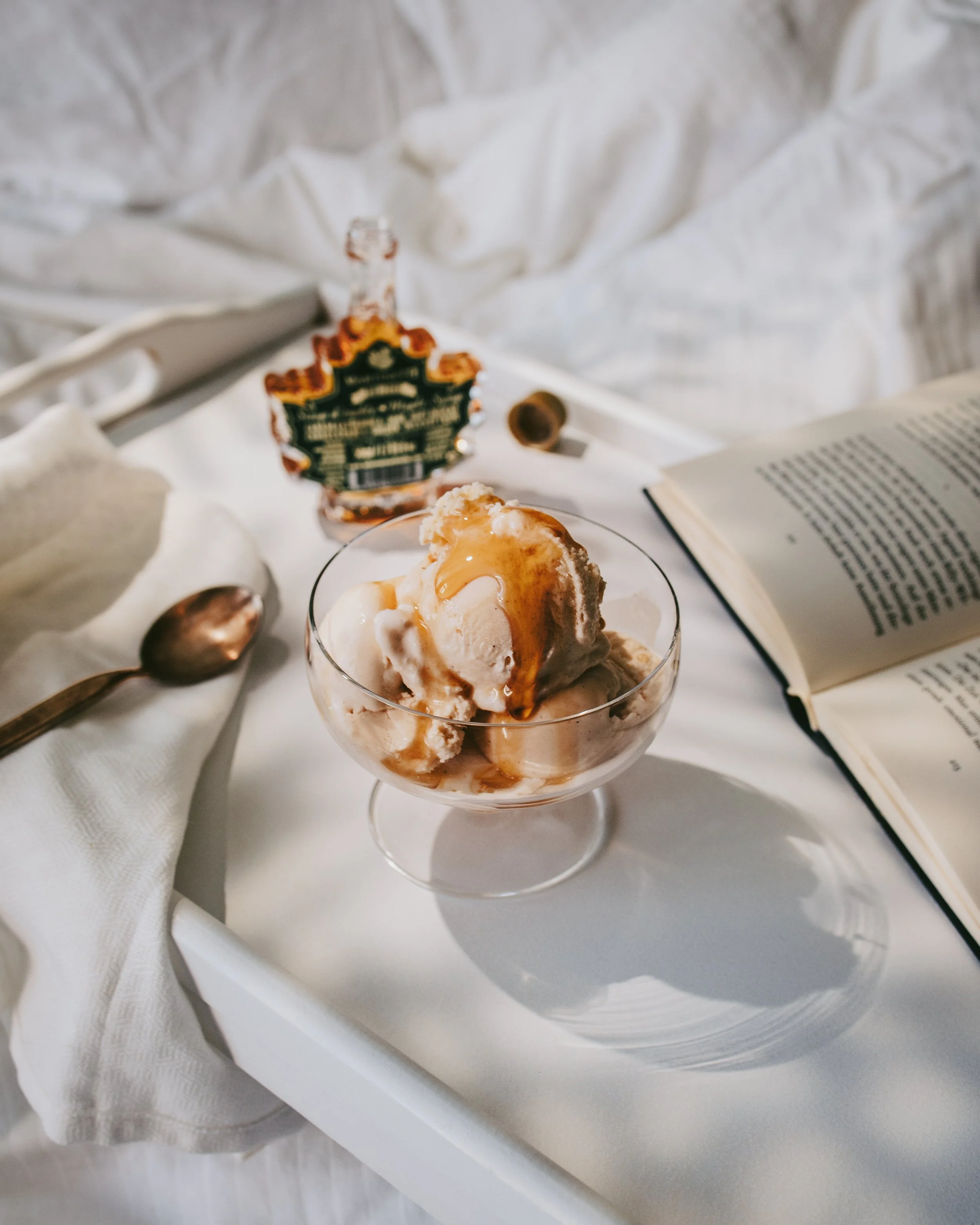 Close-up of a dessert in a glass with ice cream, caramel syrup, and whipped cream, on a white tablecloth with an open book, a spoon, and a decorative flower in the background.