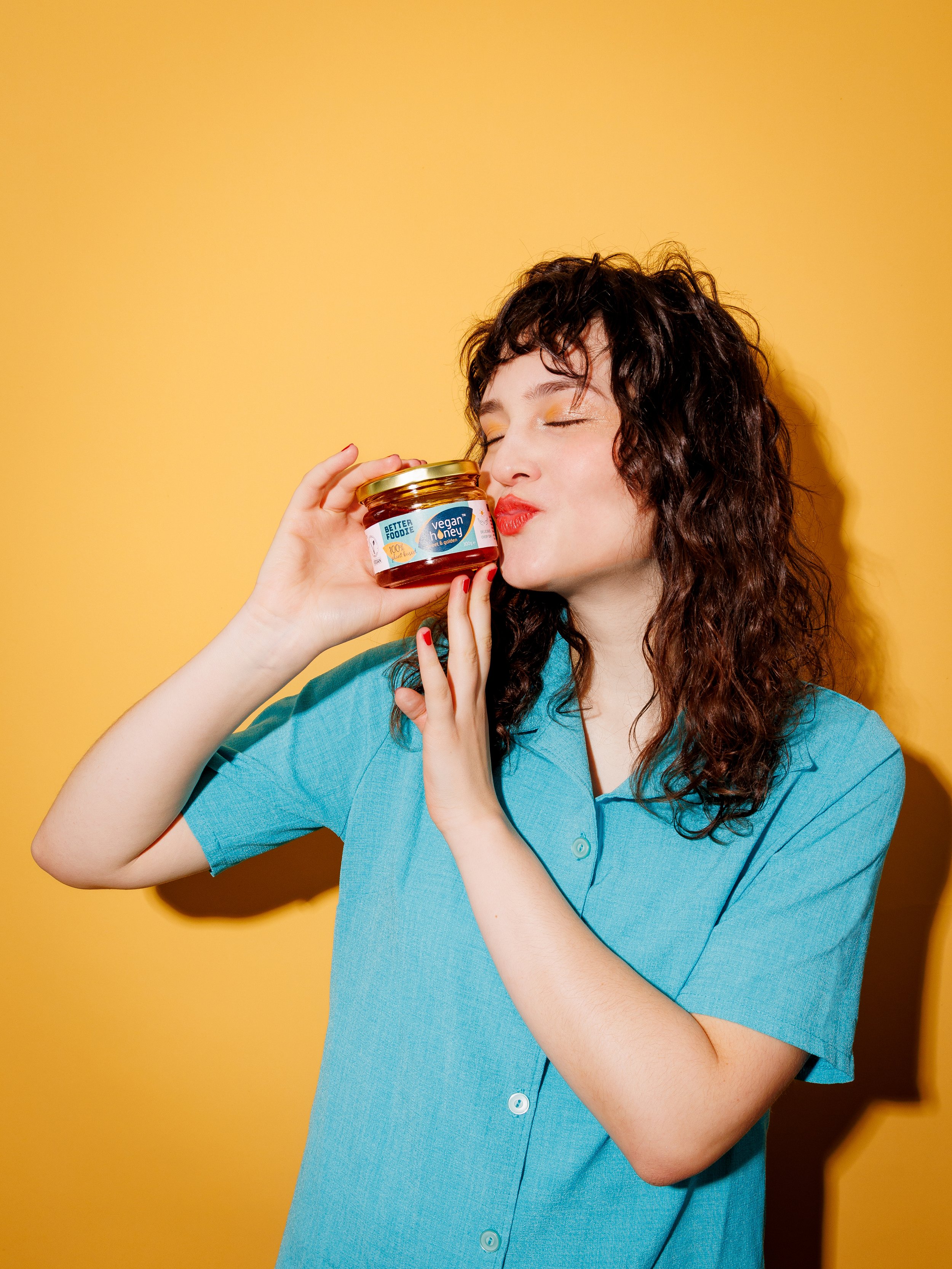 A woman with curly brown hair wearing a blue shirt holding a jar of vegan honey close to her face with her eyes closed against a yellow background.