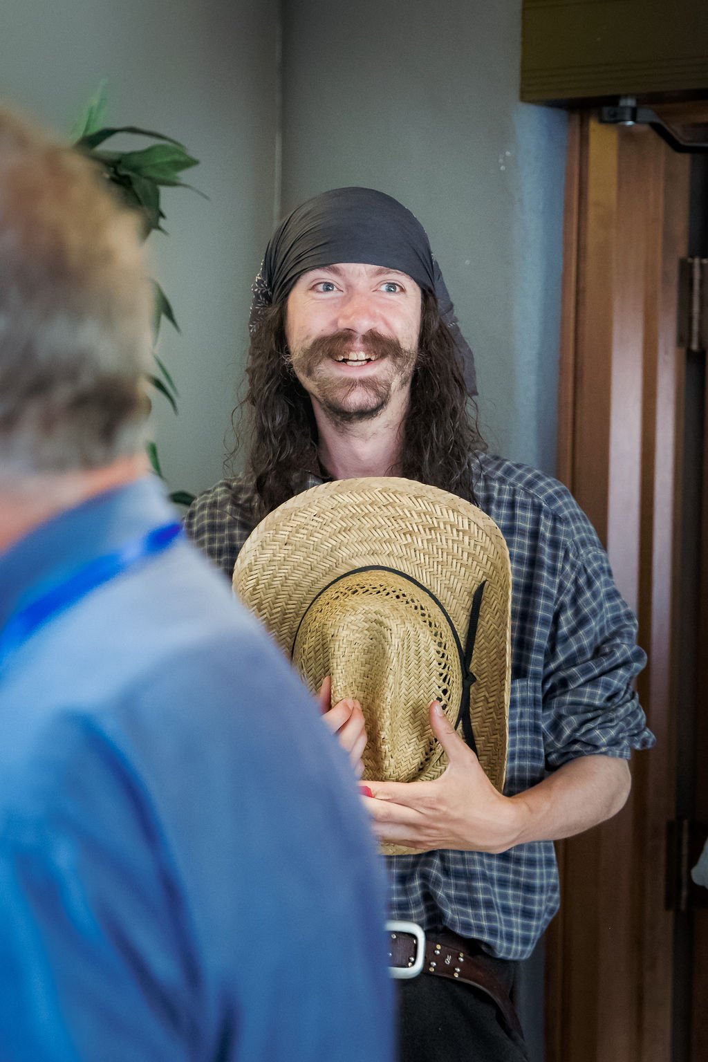 A man with long hair, a beard, and a dark bandana smiling while holding a straw hat, standing indoors near a wooden door and a green plant enclosure.