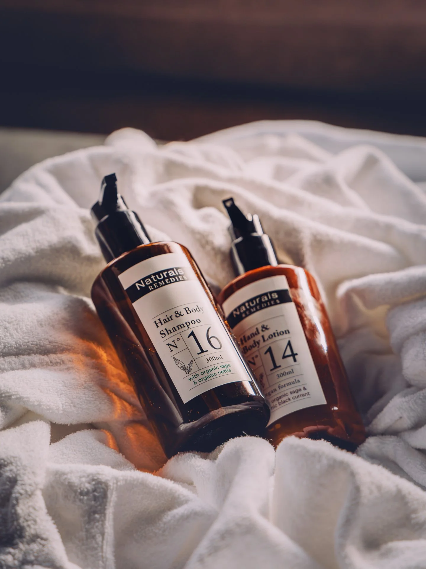 Two amber bottles of Naturals Remedies hair and body care products resting on white fabric with a dark blurred background.