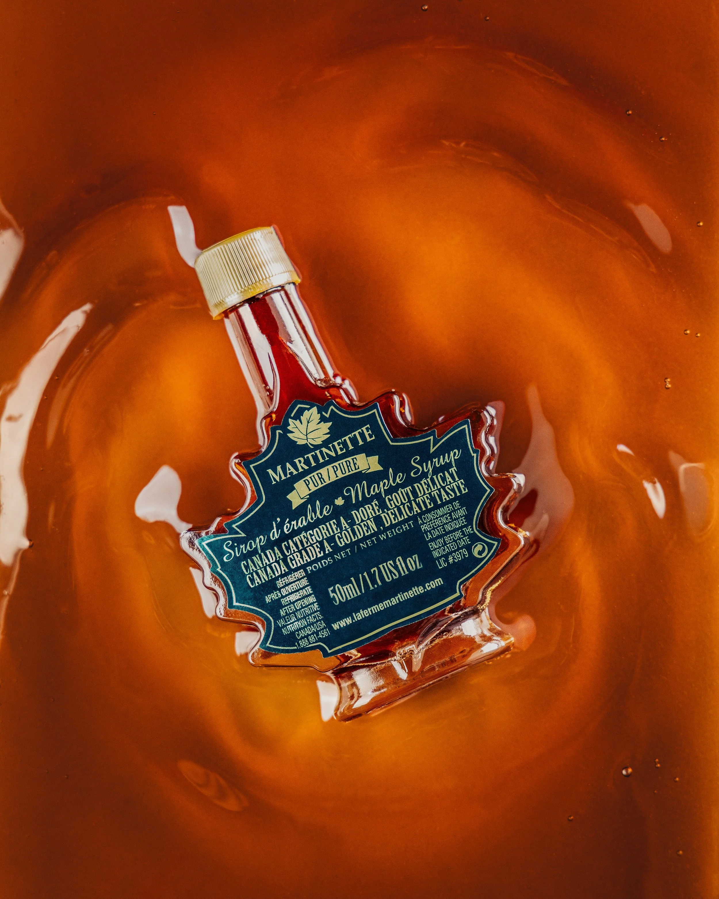 A bottle of maple syrup submerged in a bowl of amber-colored liquid, possibly syrup or caramel, with a shiny surface and some bubbles.