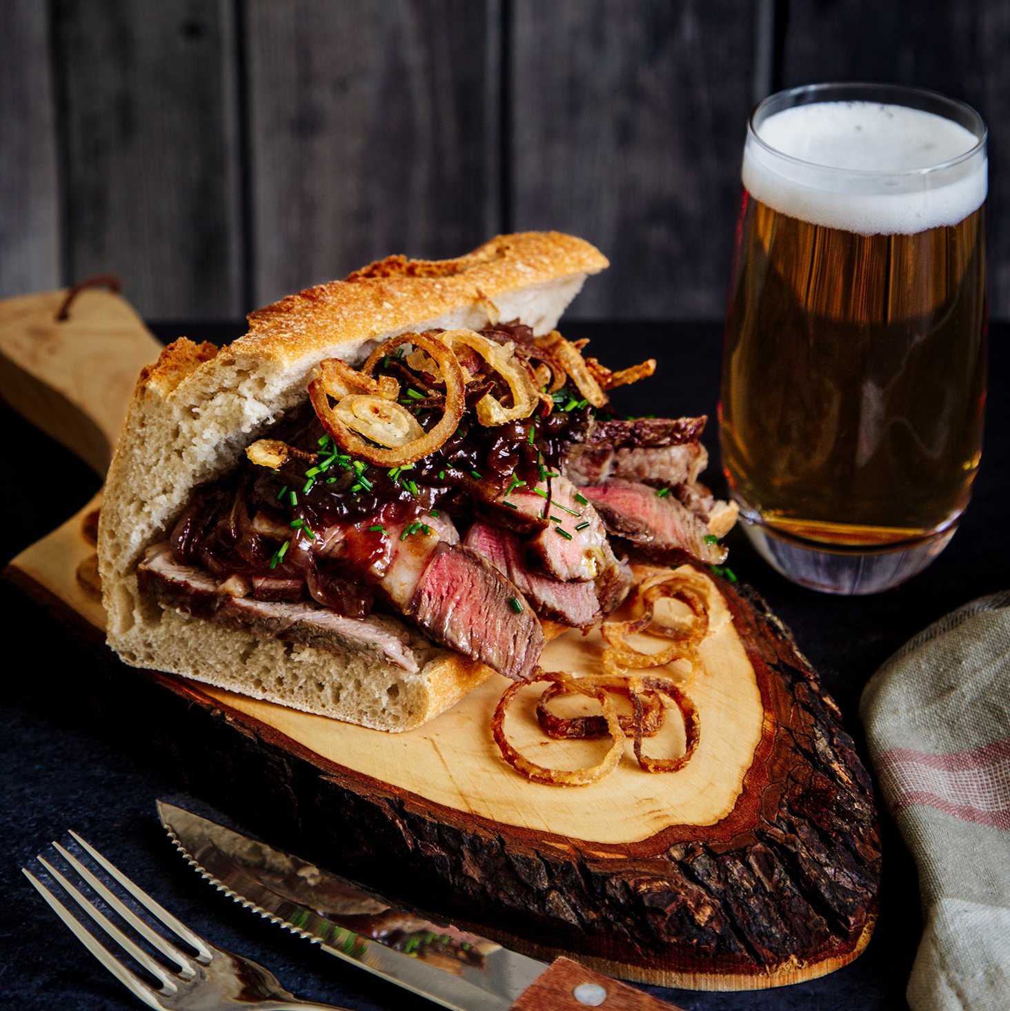 A sandwich with sliced beef, caramelized onions, and chives on a rustic wooden board, with a glass of beer beside it.