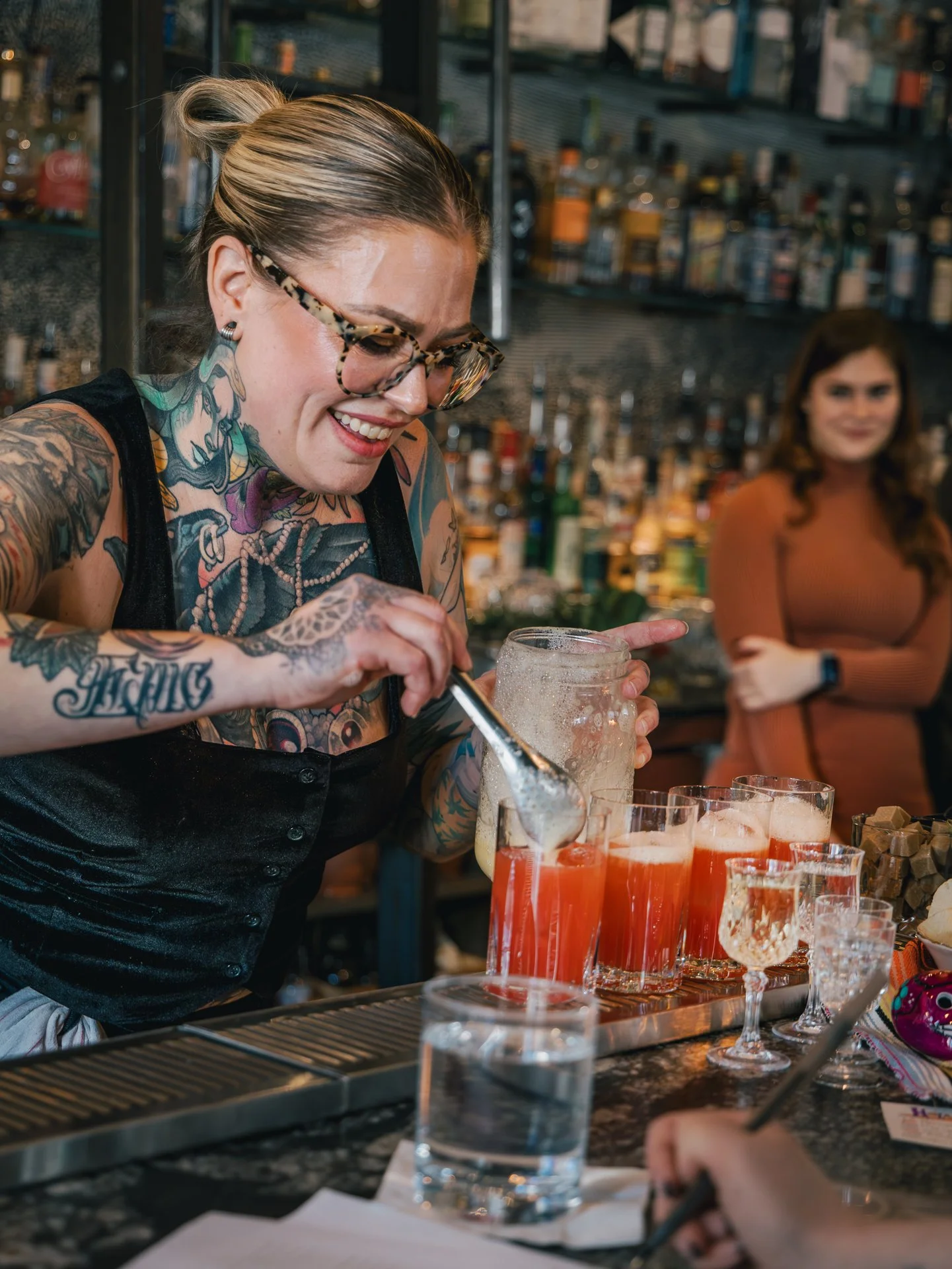 A tattooed bartender with glasses preparing drinks at a bar, with multiple glasses of red-colored beverages on the counter, and a woman in an orange-brown dress observing in the background.