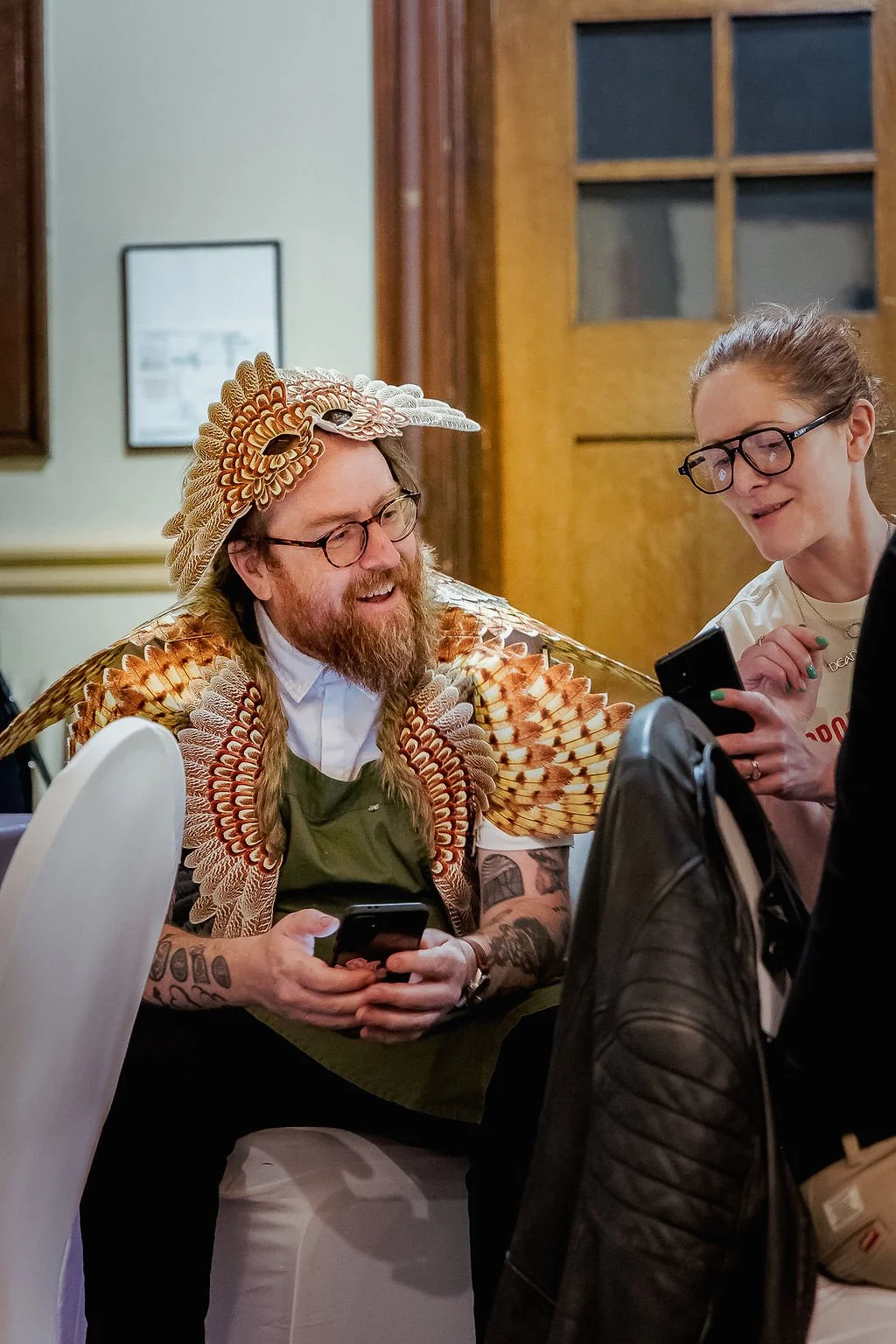 Two people, a man wearing glasses and a woman, are looking at a phone together at a table. The man is dressed in a costume resembling a bird with detailed feathers and a headpiece, and has a beard and tattoos. They appear to be indoors at a social ev