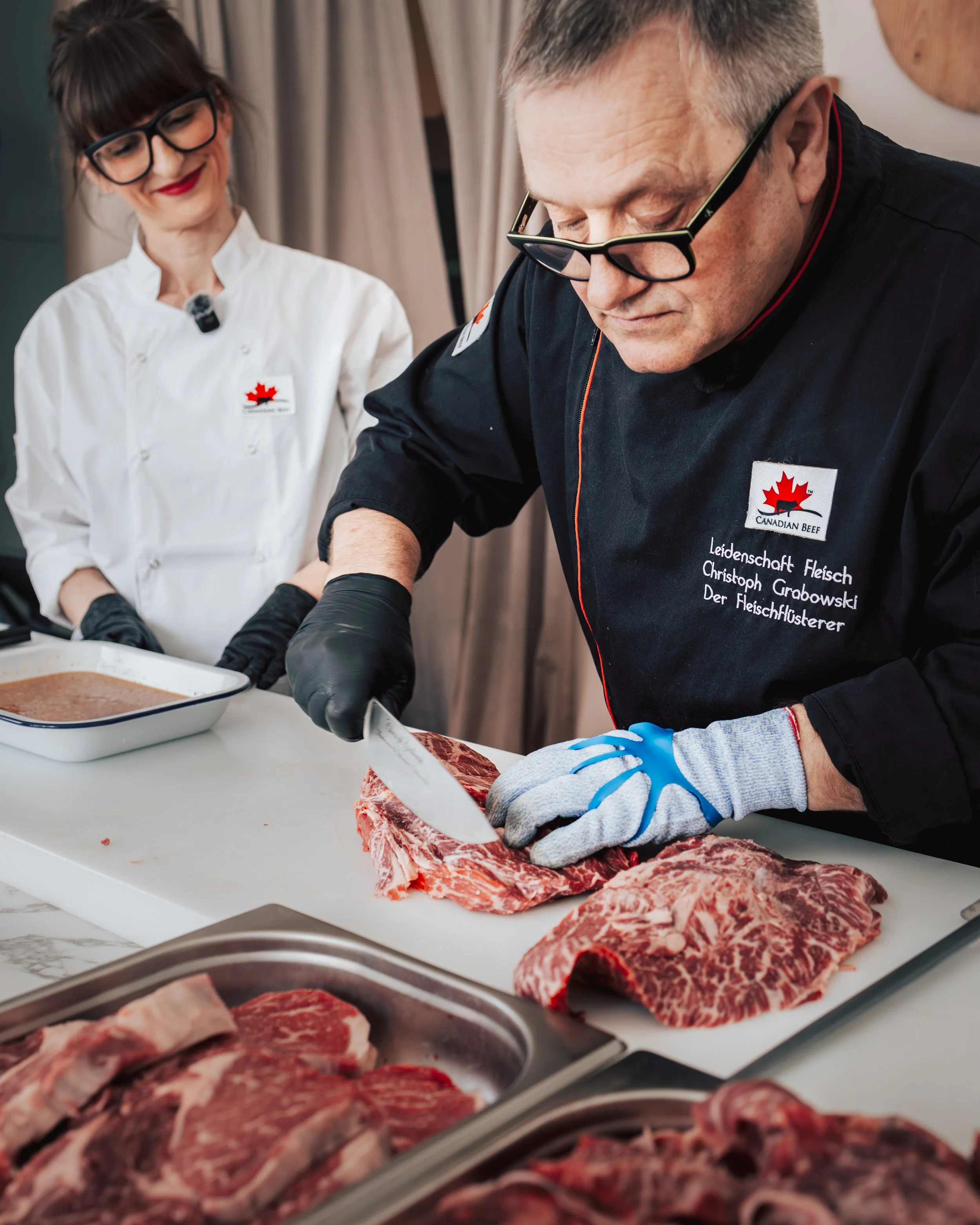 Meat being prepared on a white cutting board with a person cutting a large piece of beef with a knife, while another person watches smiling.