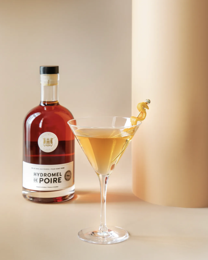 A cocktail in a martini glass garnished with a lemon twist, next to a bottle of pear-flavored spirit labeled 'Hydromel de Poire' and a beige backdrop.