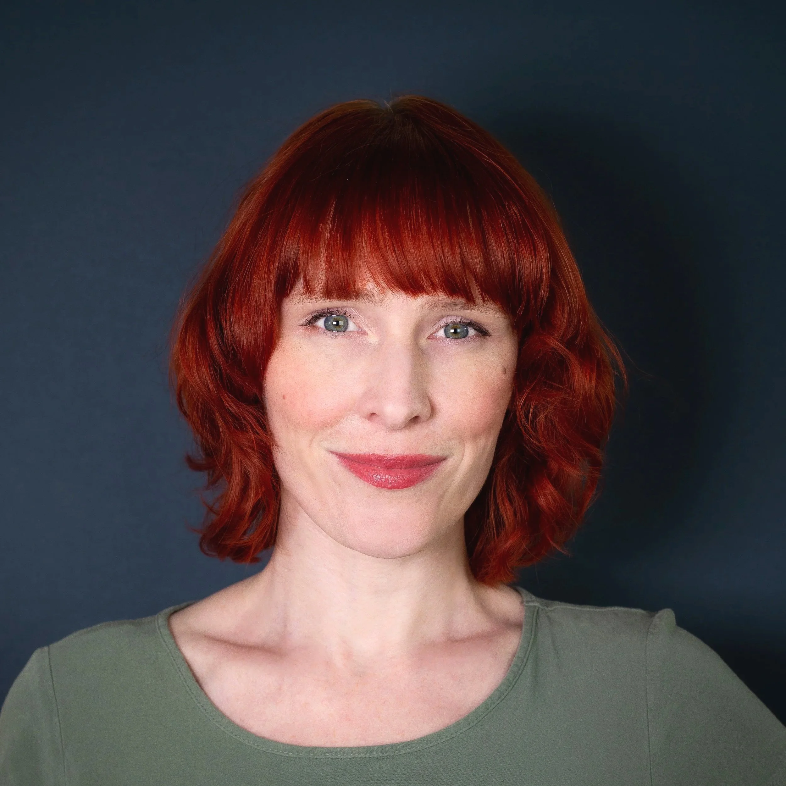 Close-up of a woman's face with curly red hair, grey eyes, and light skin, wearing a green top, with a dark background.
