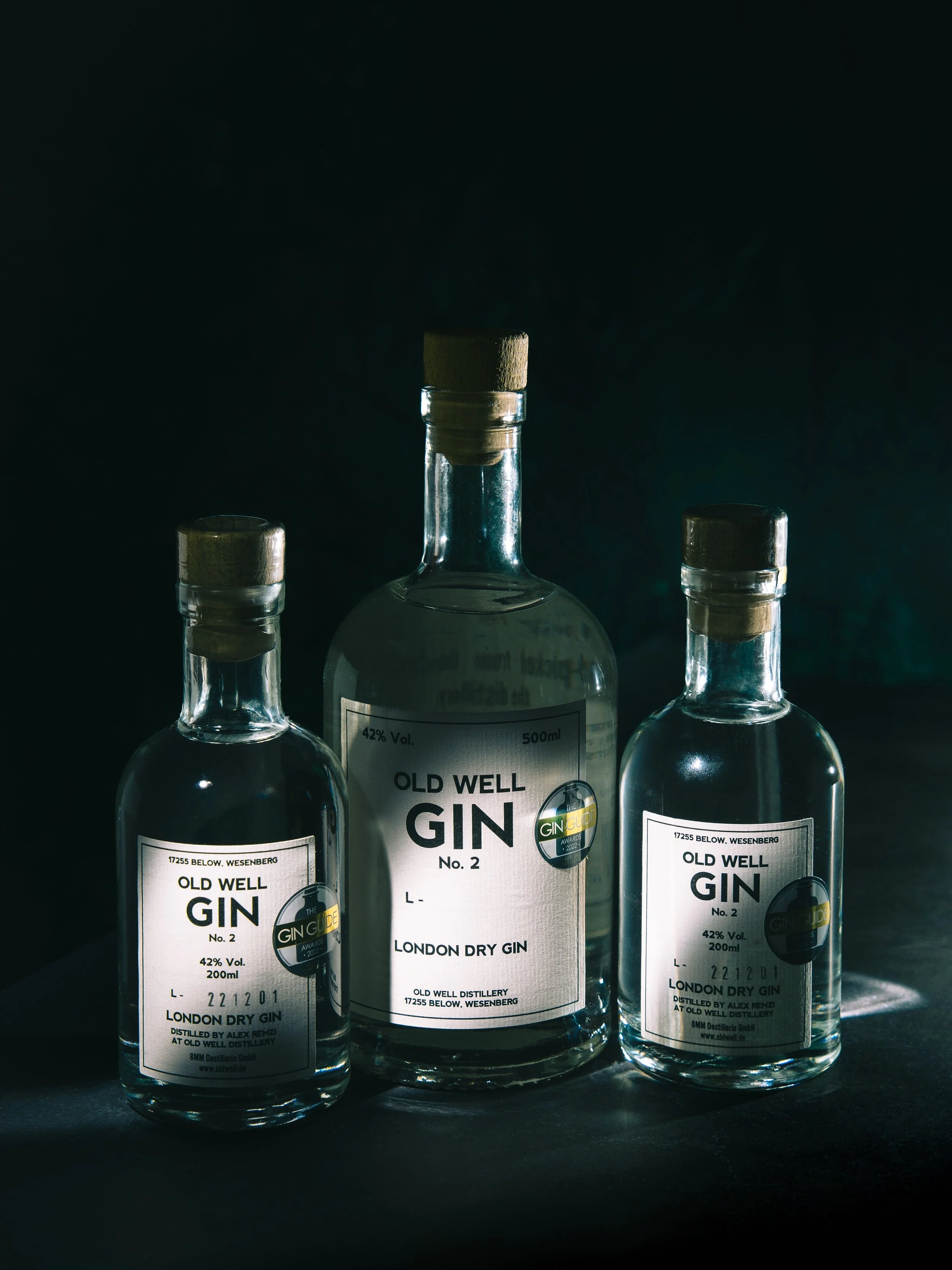 Three bottles of Old Well gin with dark background, the middle one is labeled London Dry Gin, 42% alcohol, 200ml, with the other two bottles partially visible on either side.