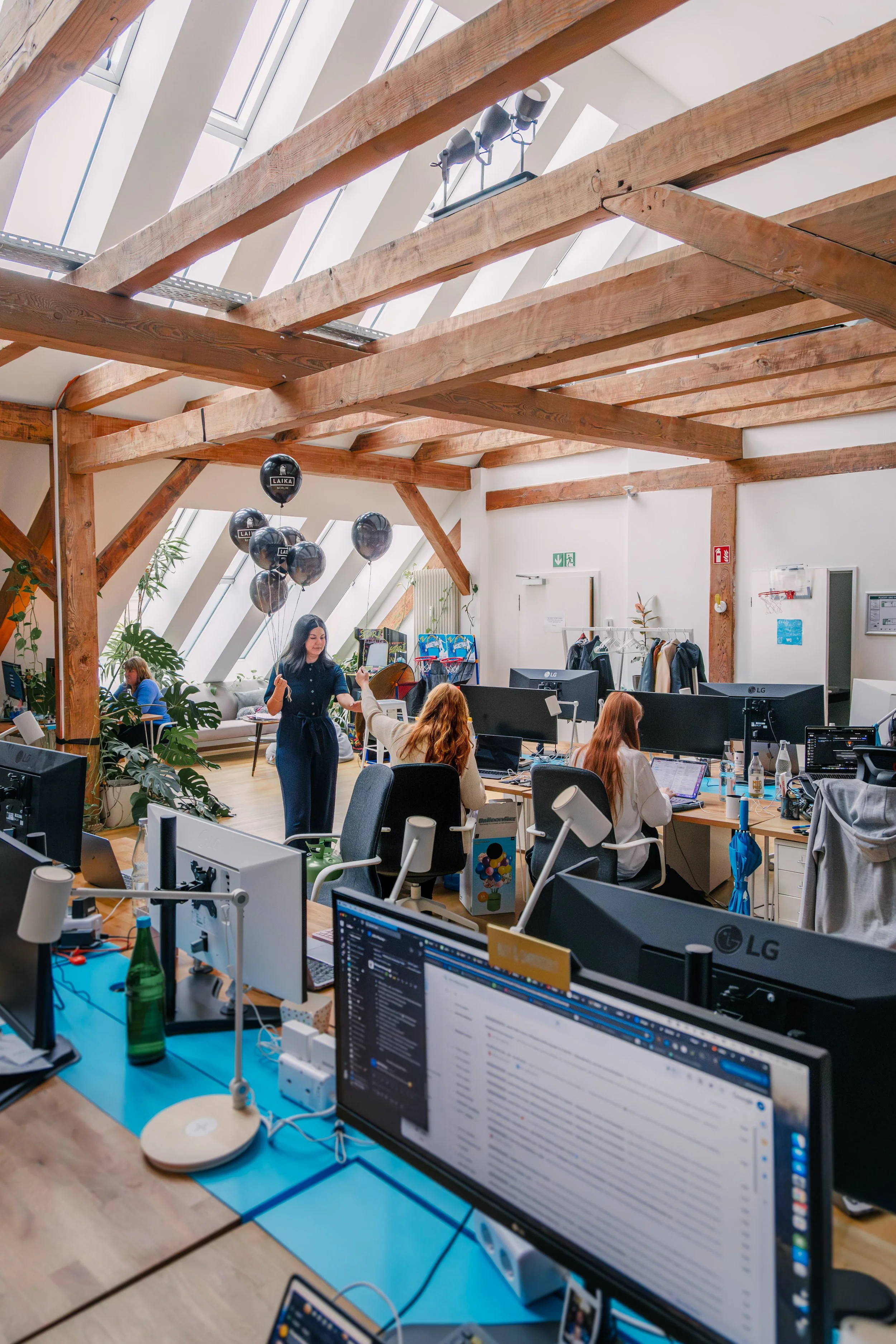 A busy modern office with multiple computer workstations, people working at their desks, and decorative elements like black balloons, plants, and light fixtures. The office has large skylights and exposed wooden beams.