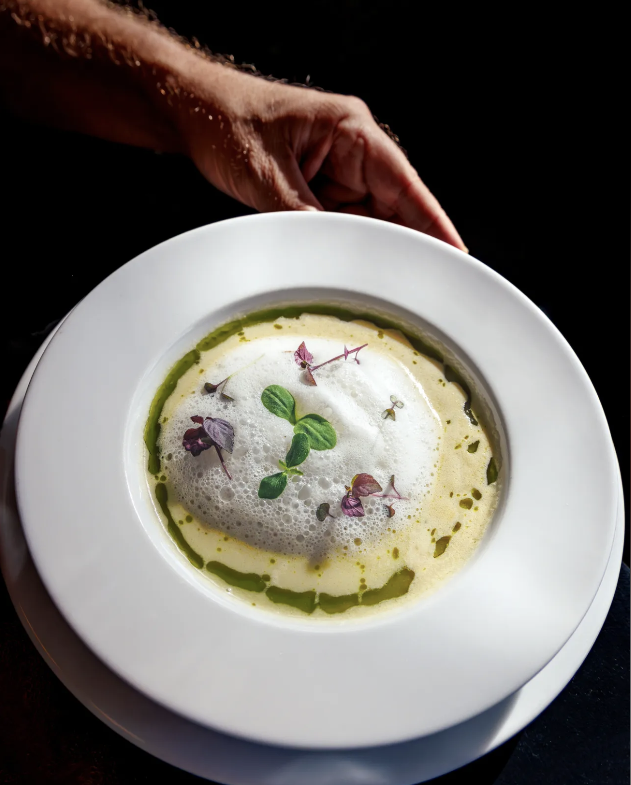 A gourmet soup garnished with microgreens and edible flowers, served in a white bowl on a dark background.