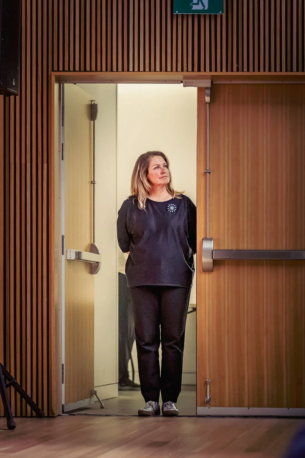 A woman with shoulder-length blond hair standing in a doorway, looking to her right, in an indoor setting with wooden paneling.
